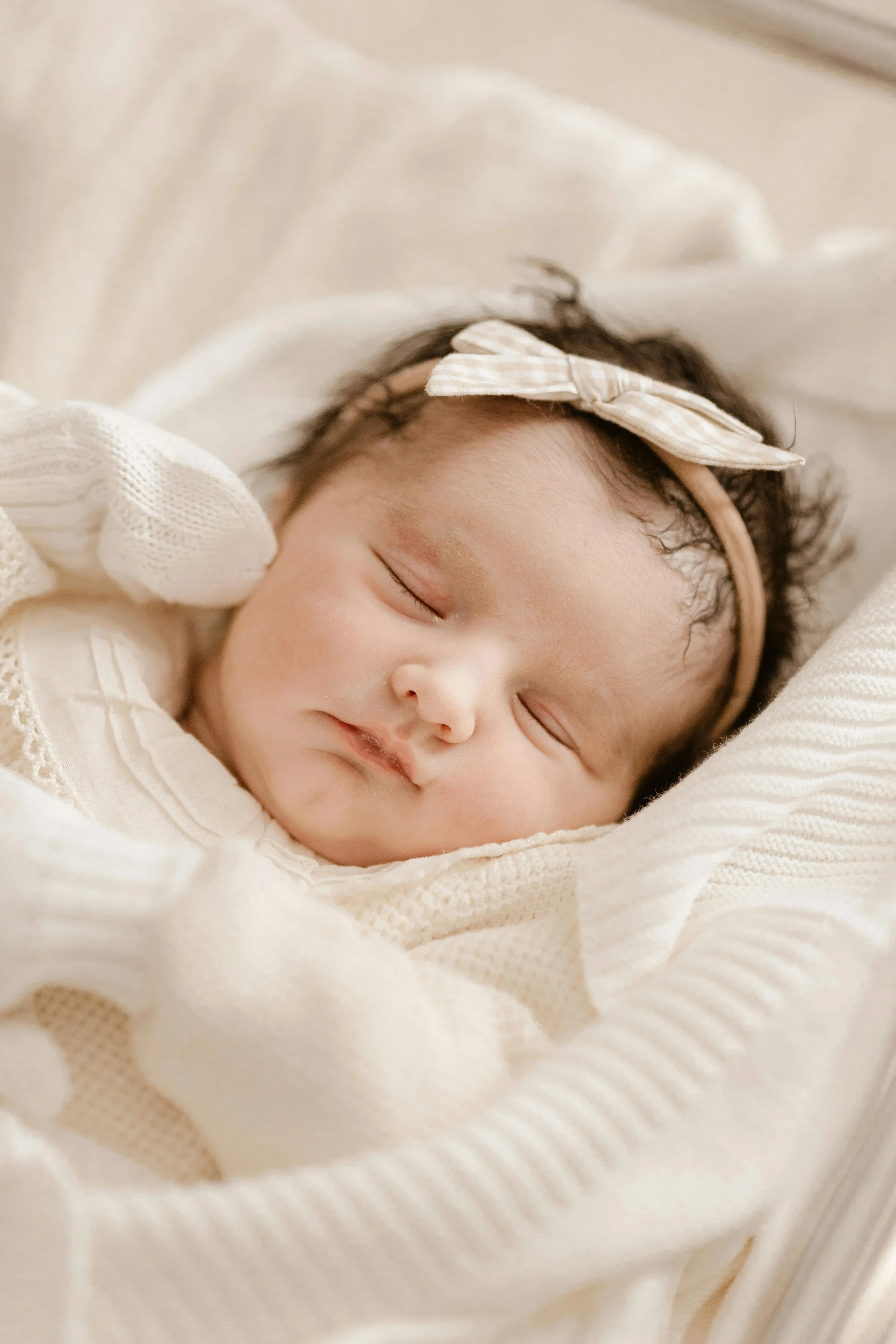 A sleeping baby lying on a soft, cream-colored blanket, wearing a cream-colored outfit with lace details and a beige headband with a bow.