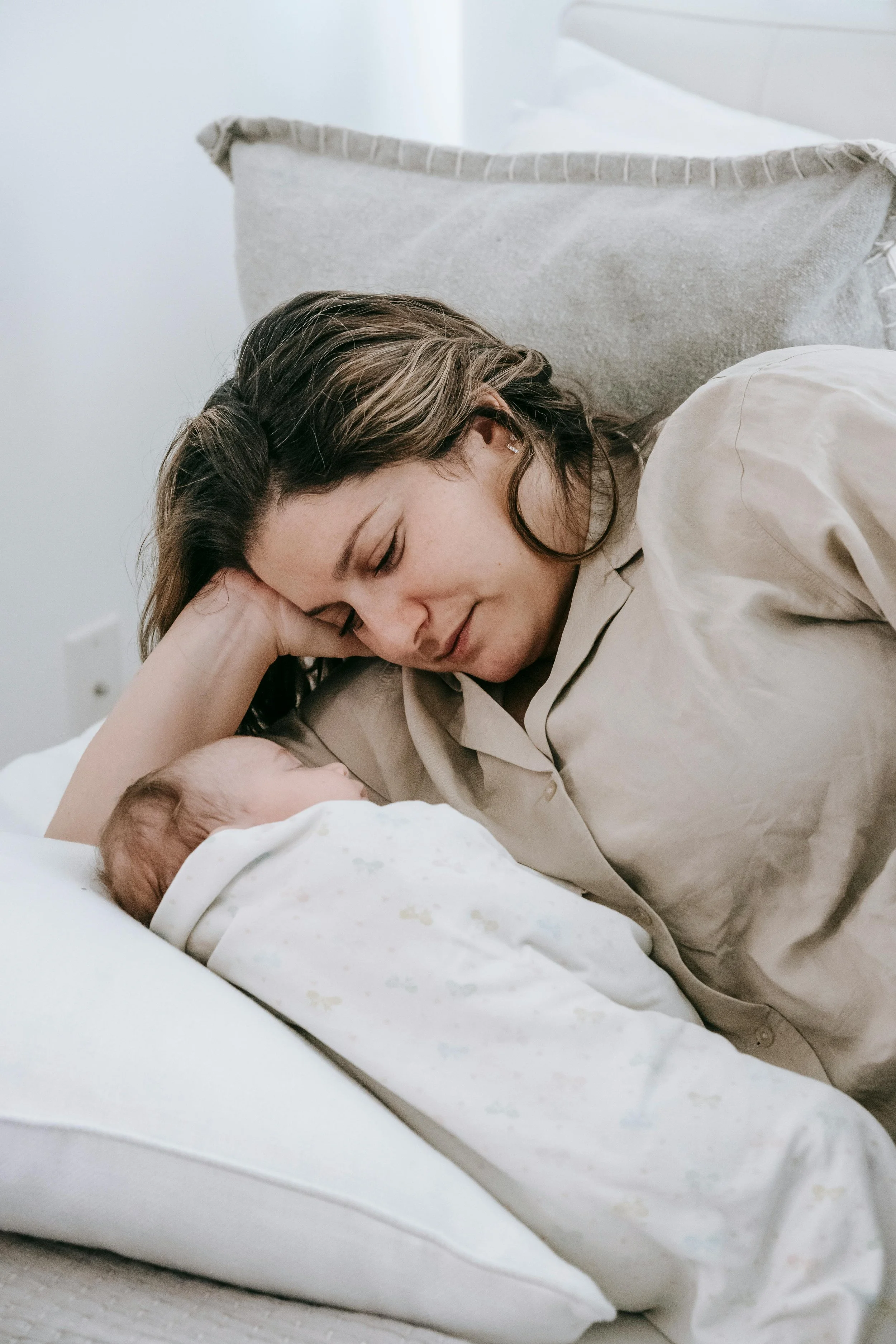 A woman with brown hair resting her head on a pillow, leaning over to look at a newborn baby lying in a hospital bassinet. The baby is wrapped in a blanket.