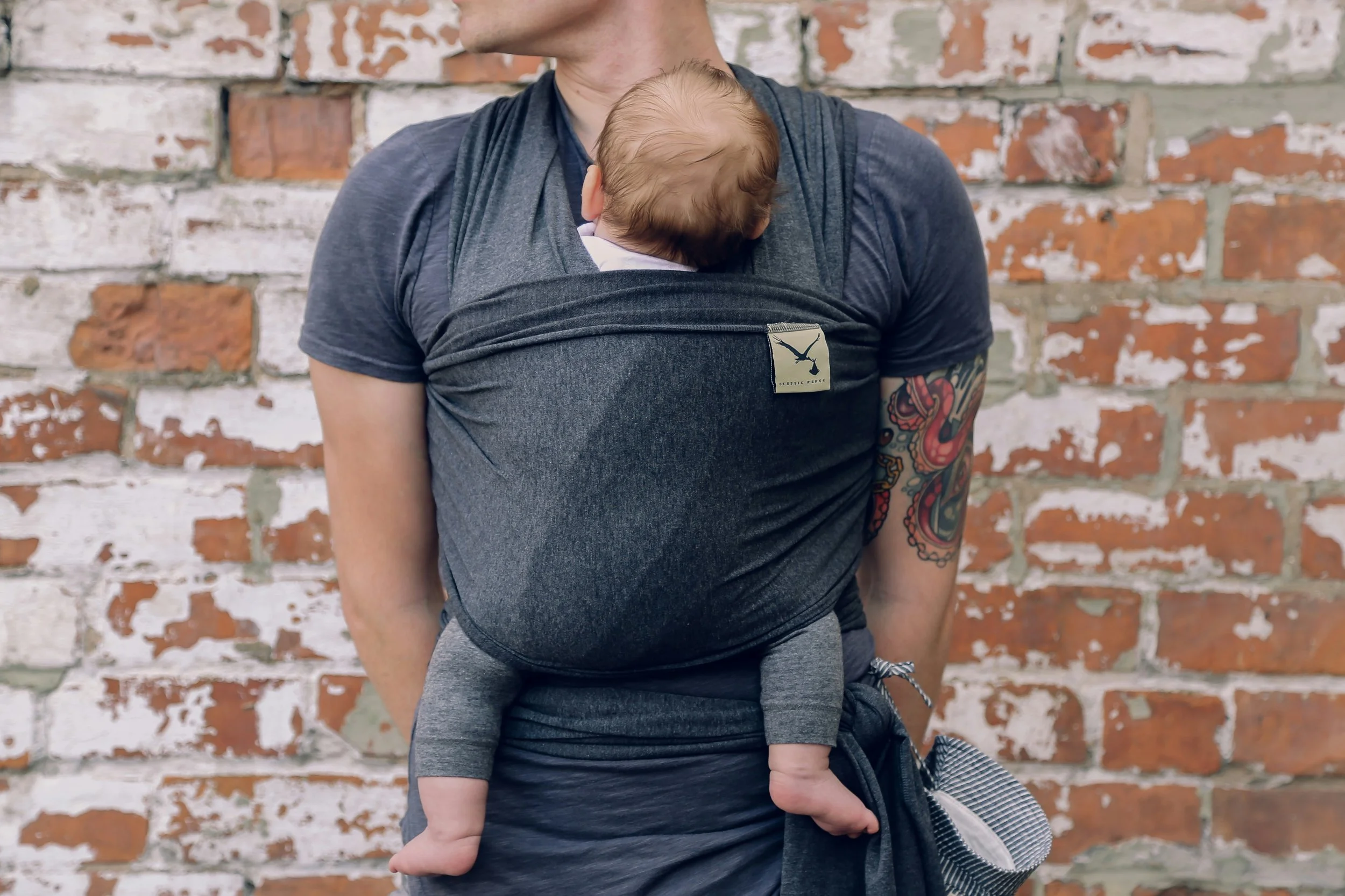 Person carrying a baby in a gray baby carrier against a weathered brick wall.
