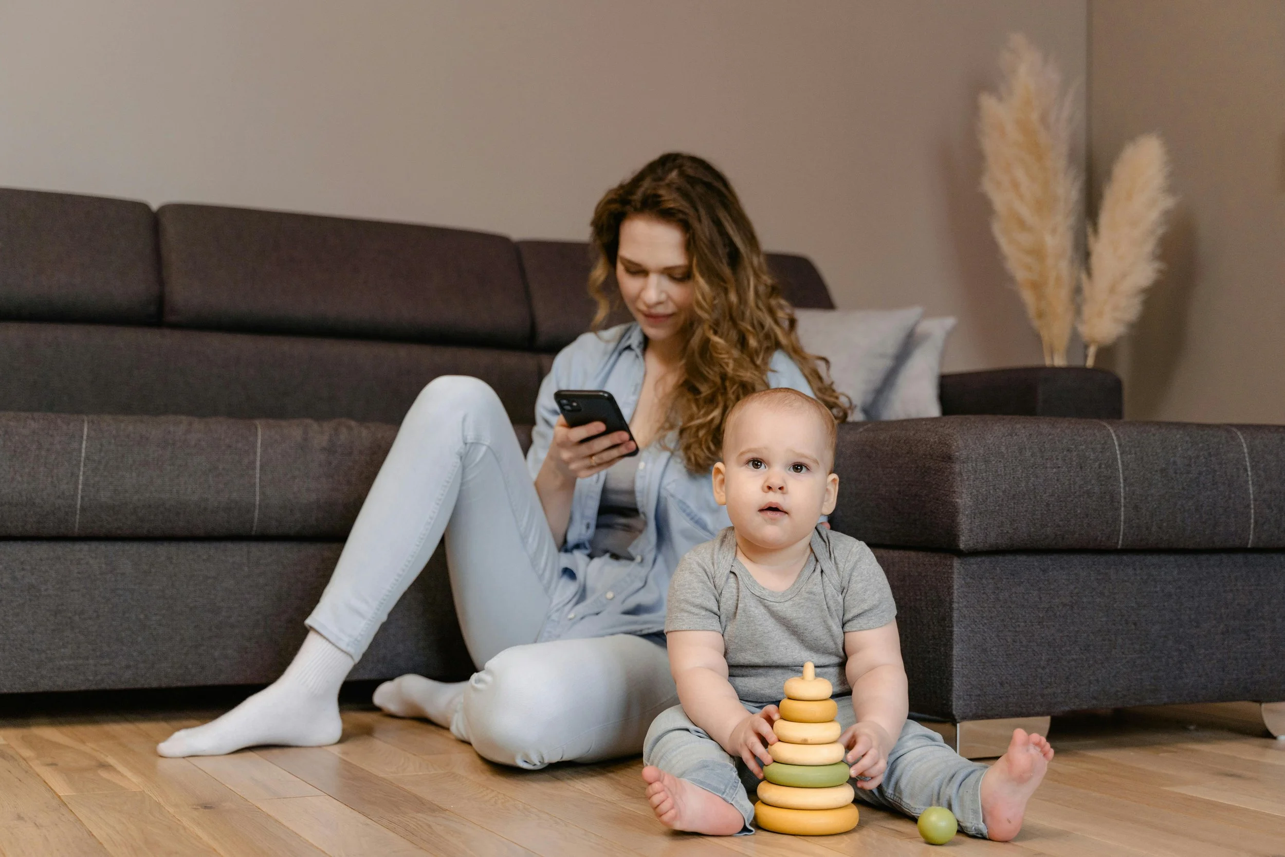 A woman with curly hair sits on the floor beside a young child, looking at her phone. The child, with a serious expression, is playing with a body of stacked wooden rings and a green ball.