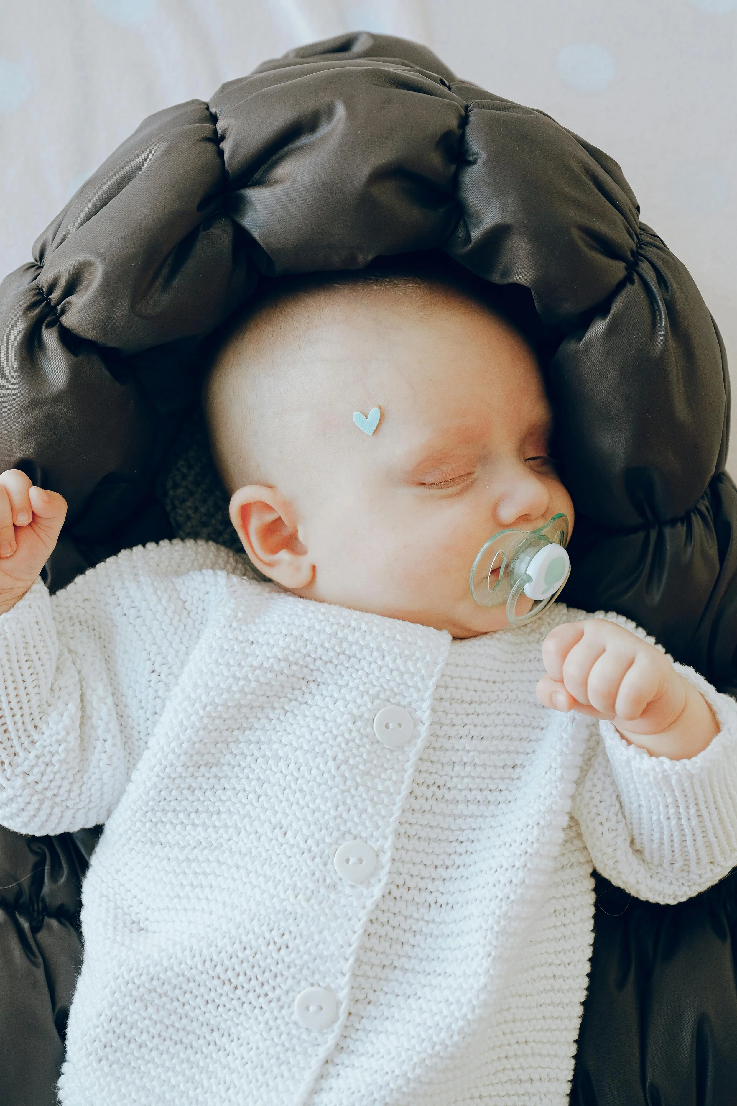 Close-up of a sleeping baby with a pacifier in their mouth, wearing a white knitted sweater, resting on a black padded surface, with a light blue heart-shaped sticker on their forehead.
