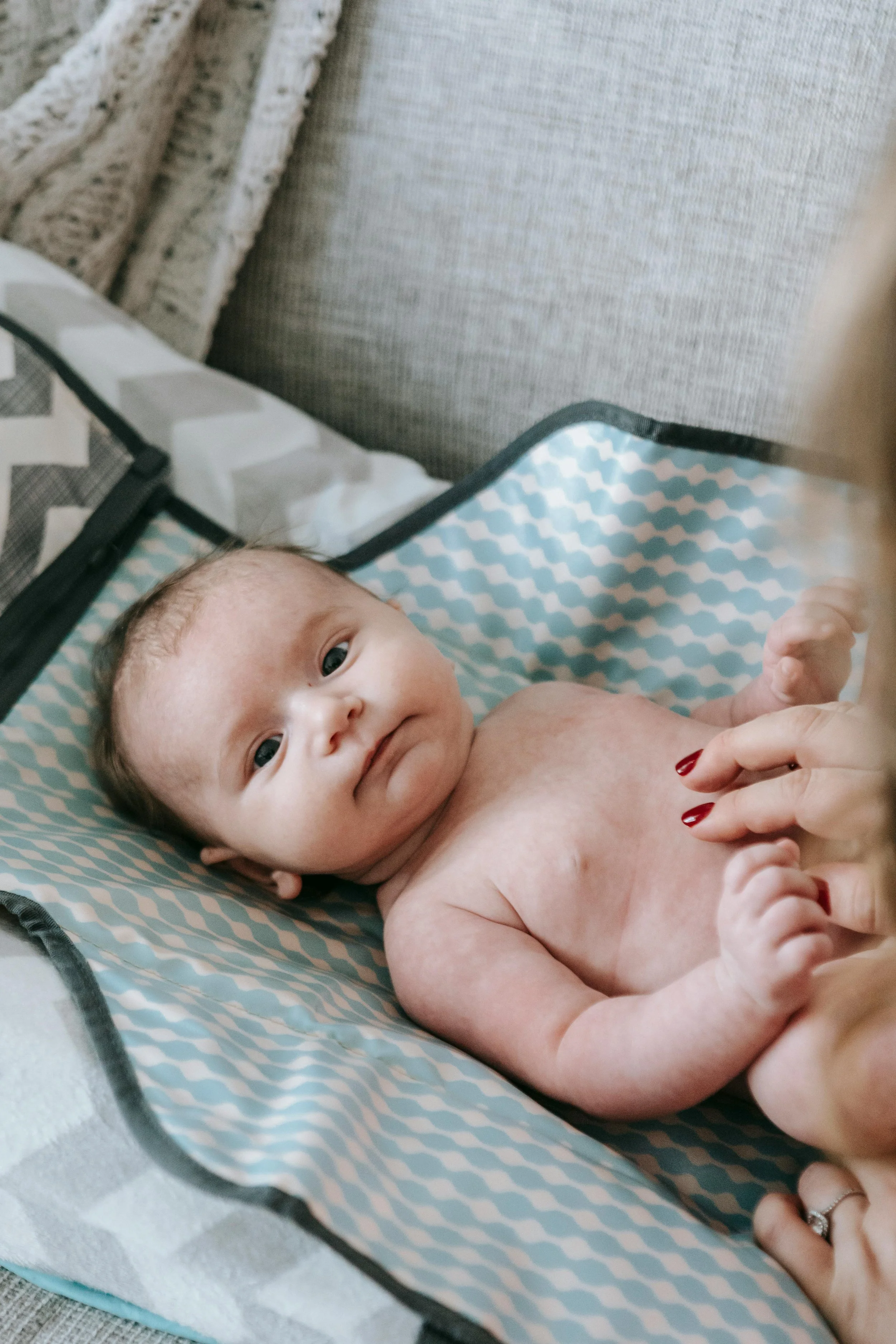 A baby lying on a blue and white patterned changing pad, looking at the camera, with an adult's hand resting nearby, wearing a ring.