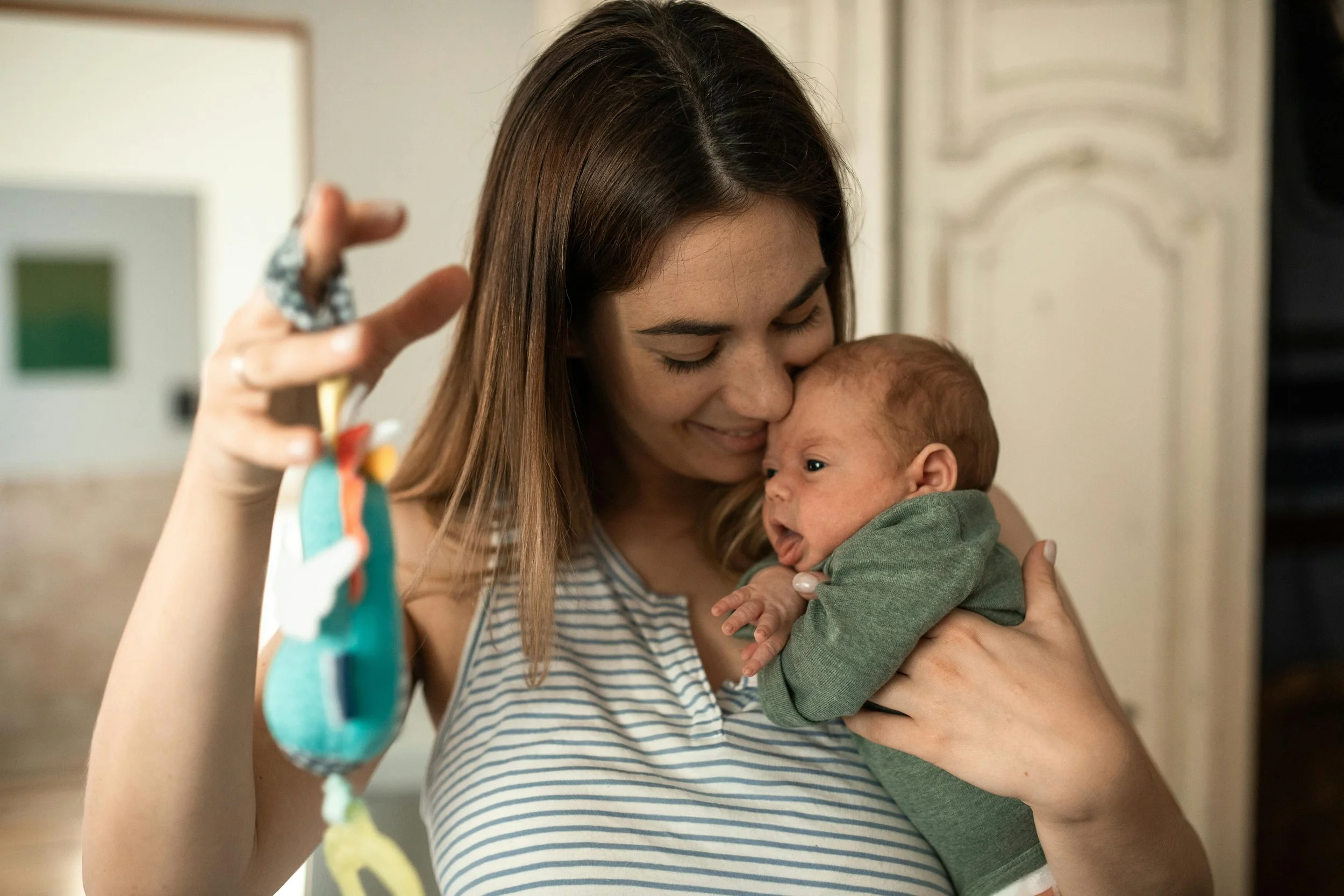 A woman with long brown hair holding a baby close, smiling and looking down at the baby. The woman is wearing a striped shirt and a ring on her finger. The woman is holding a colorful, soft toy in her right hand.