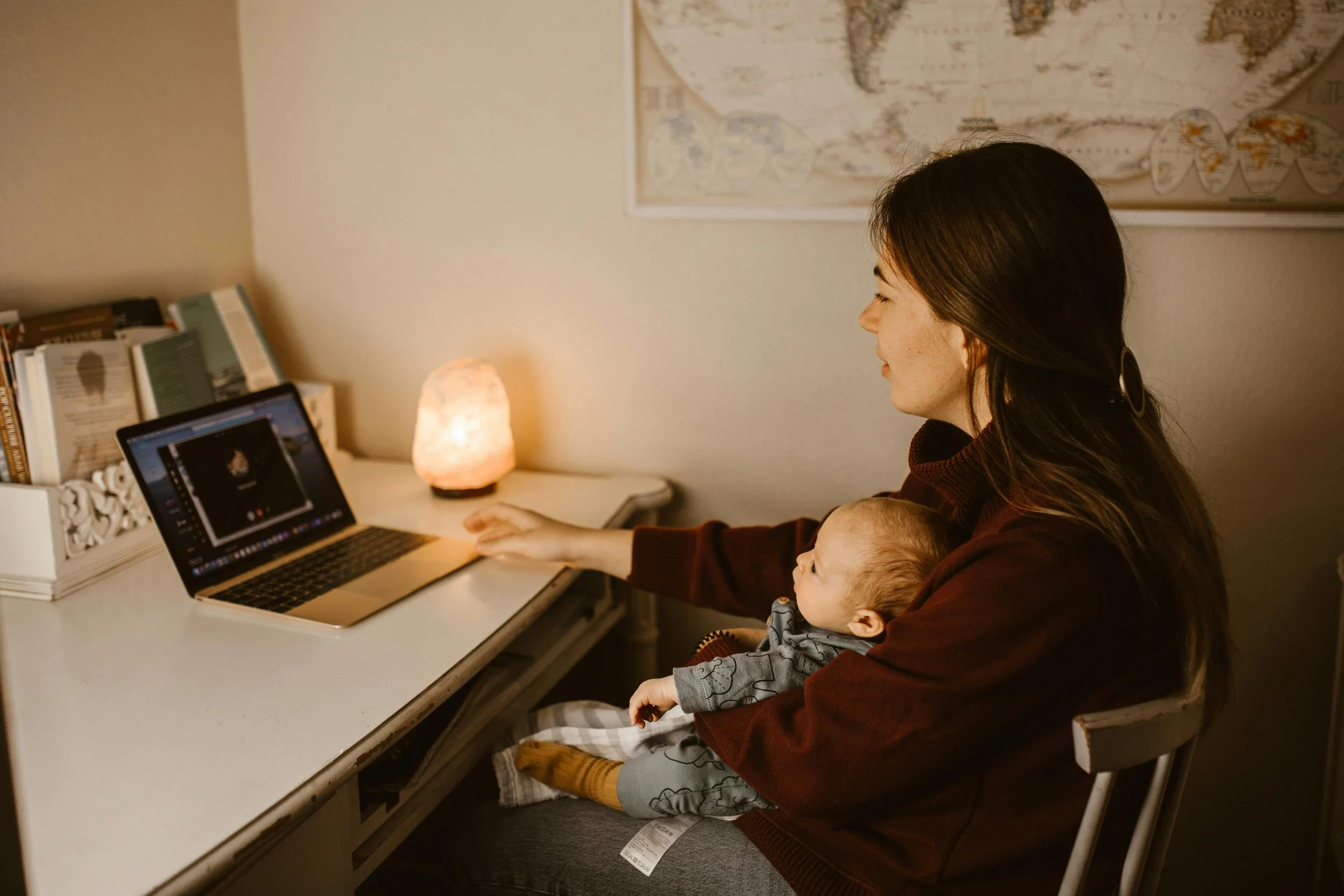 A woman sitting at a desk with a baby on her lap, looking at a laptop, in a dimly lit room with books and a salt lamp on the desk, and a world map on the wall behind.