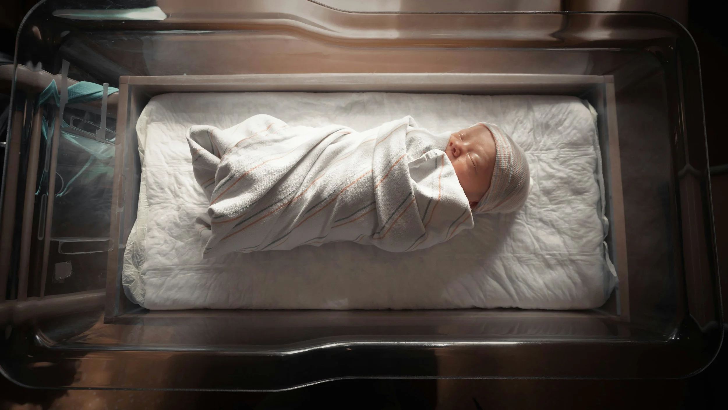 A newborn baby wrapped in a striped blanket and wearing a knit hat, lying in a clear hospital bassinet.