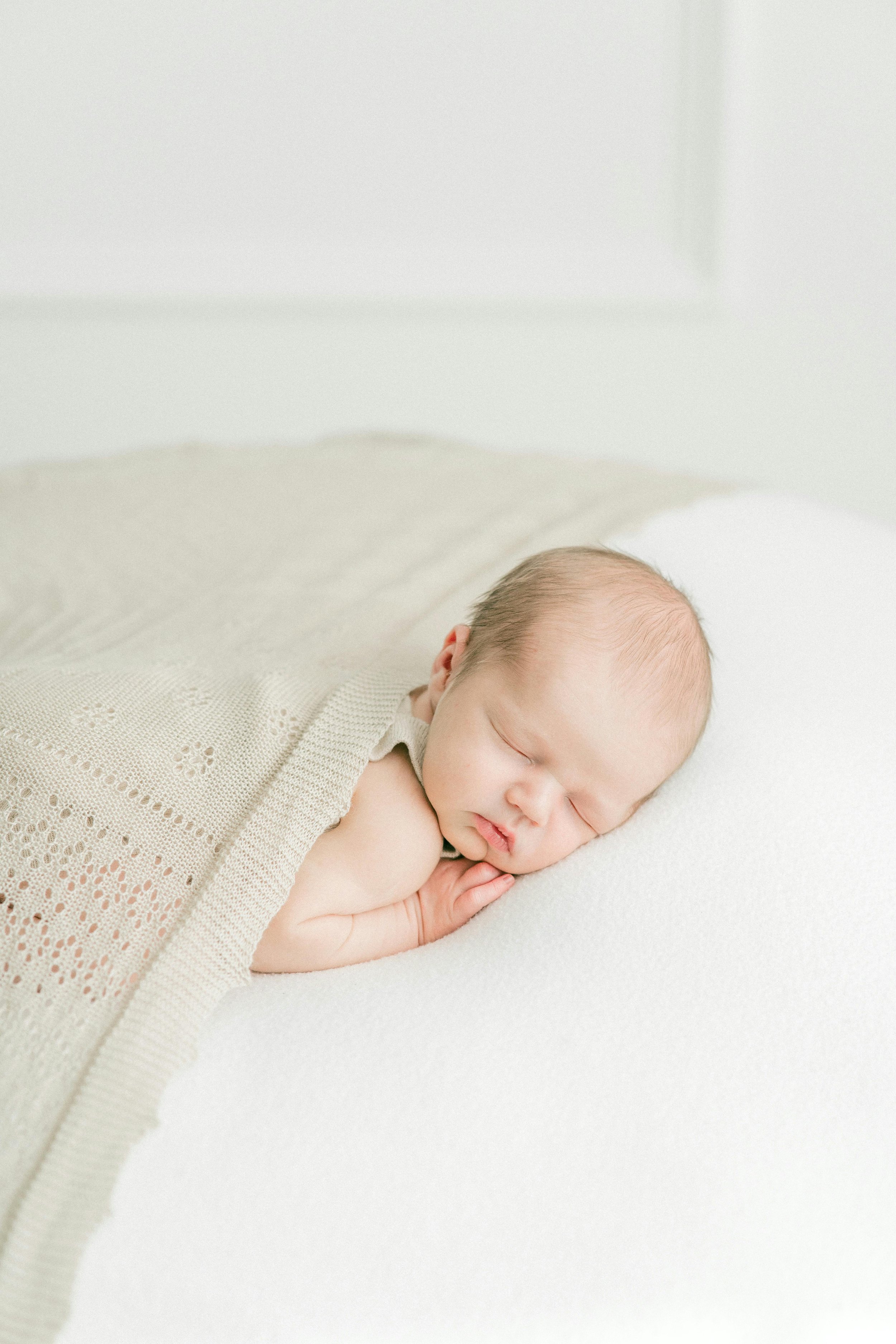 A sleeping newborn baby with a beige knitted blanket on a white surface.