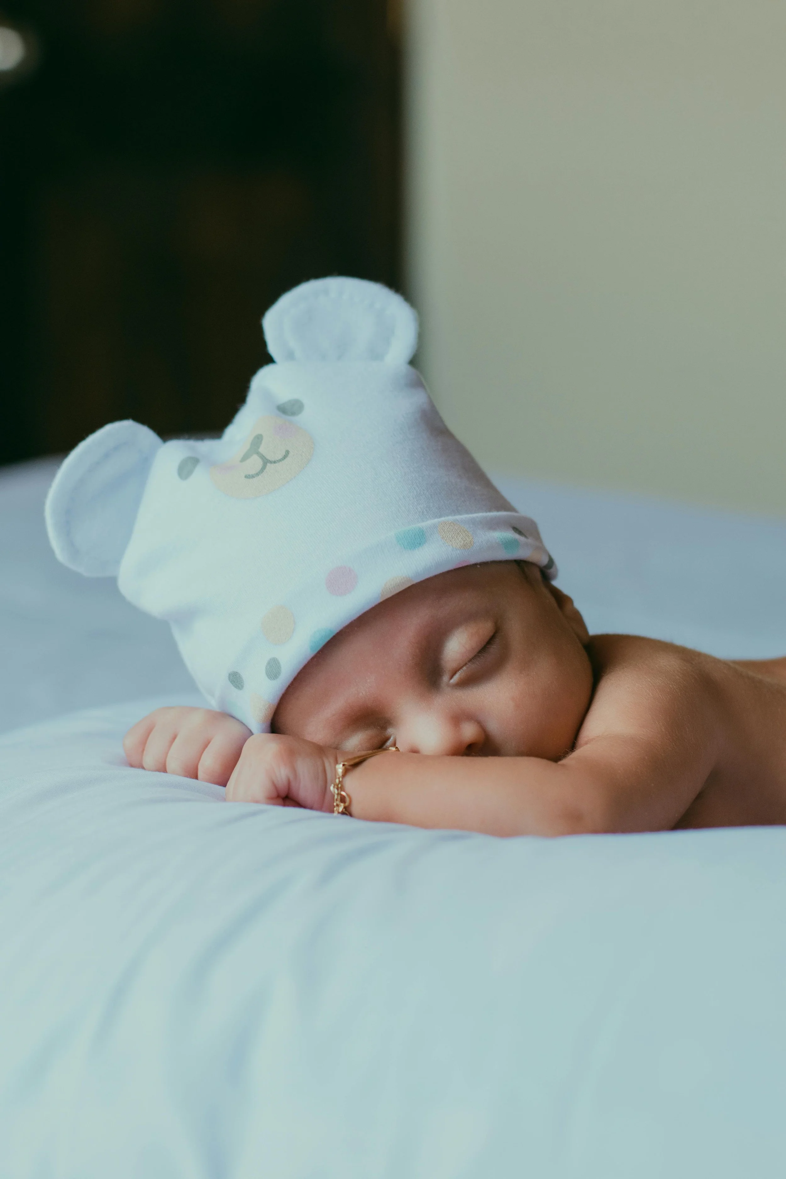 A sleeping newborn baby lying on a white surface, wearing a white hat with bear ears and a pastel polka dot pattern.
