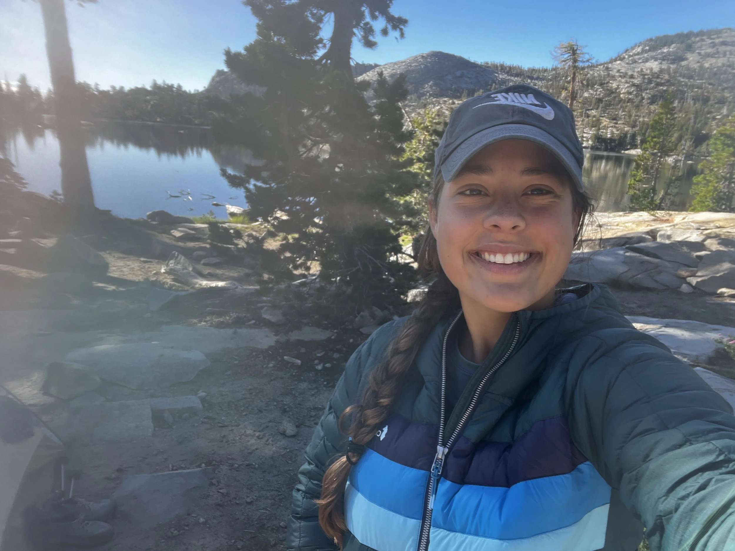 A woman smiling while taking a selfie outdoors near a lake with trees and mountains in the background.