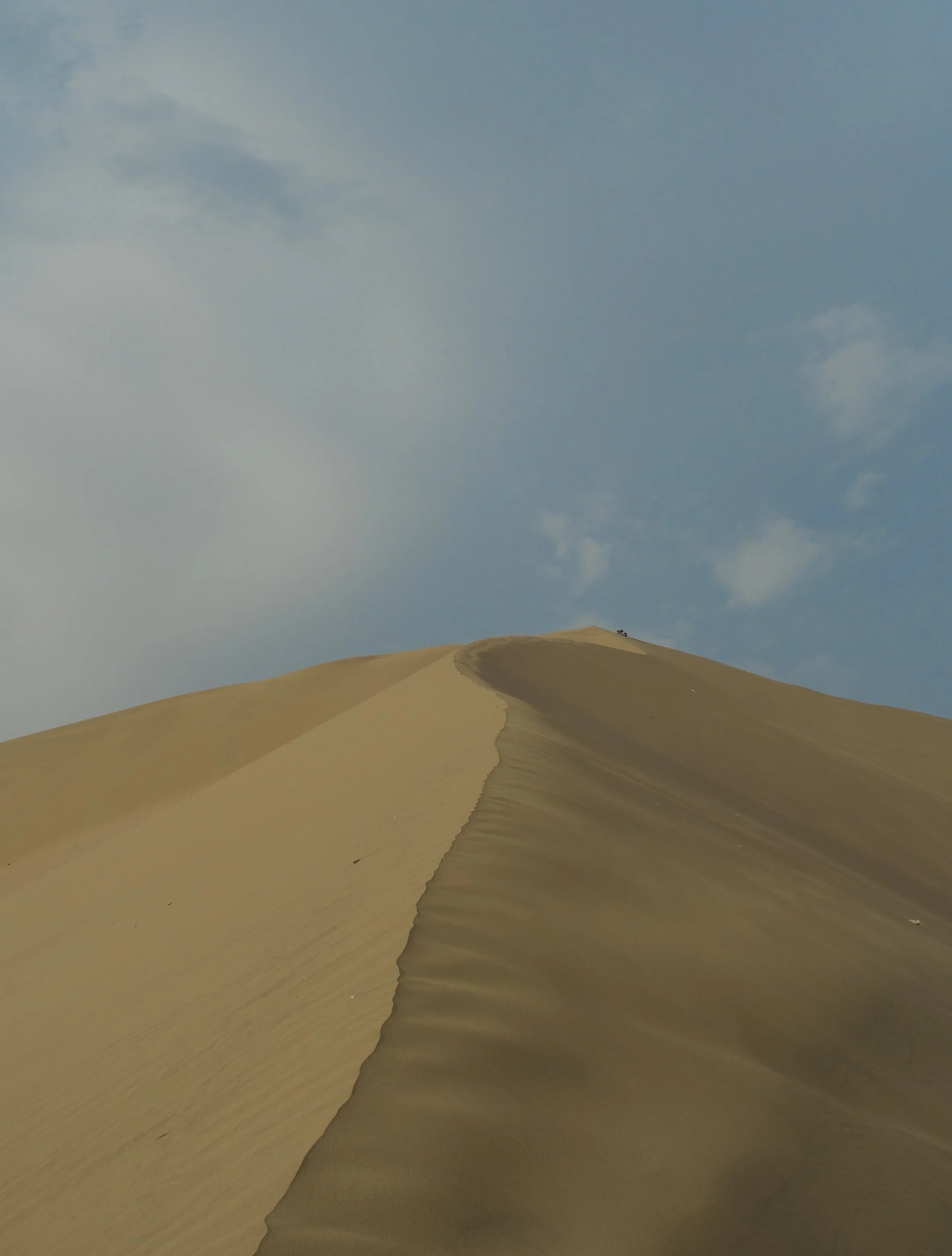 Dune in a desert with a few people at the top, under a partly cloudy sky.