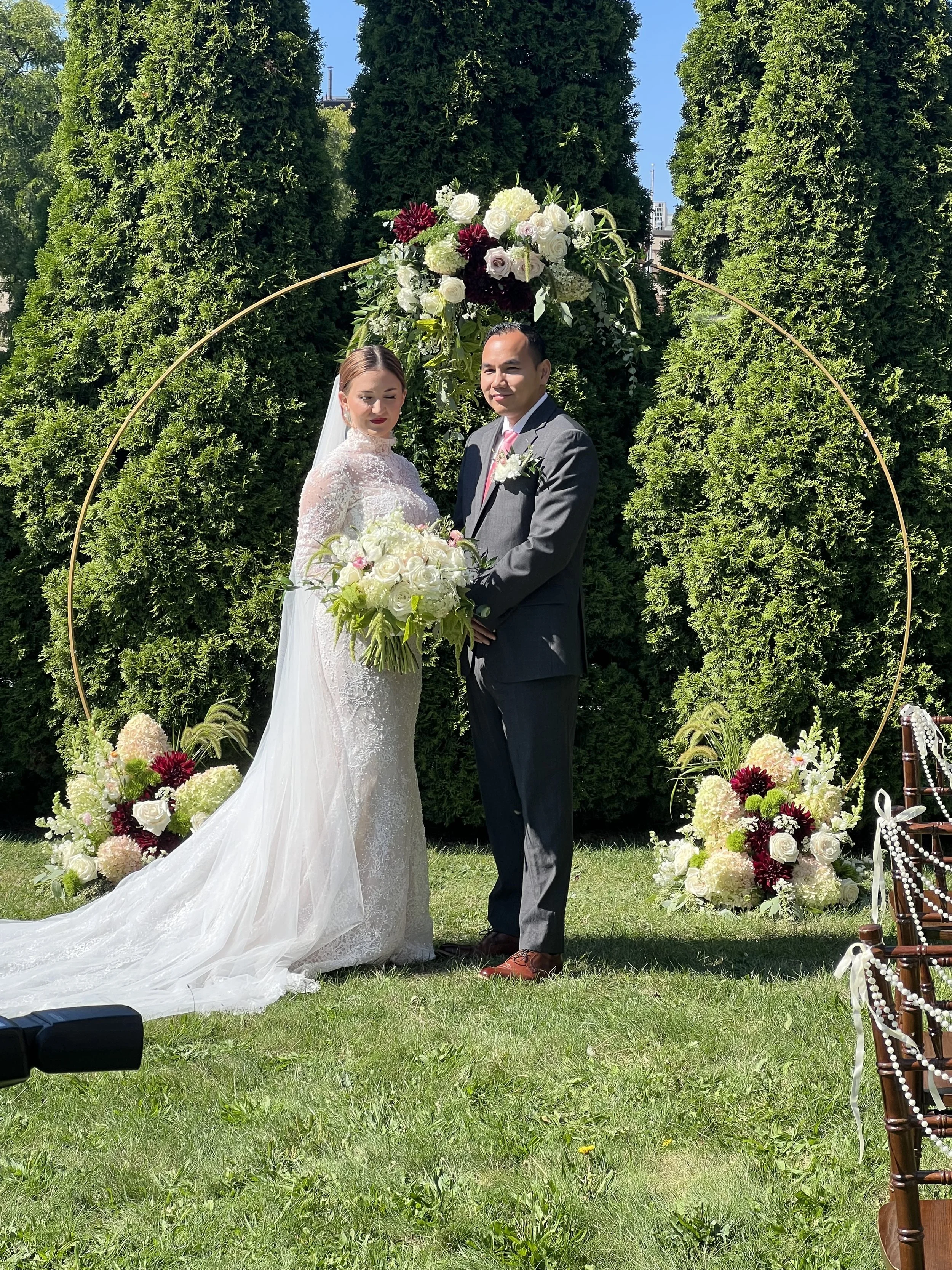 A bride and groom standing beneath a floral arch, holding hands and smiling at the camera, in an outdoor wedding ceremony setting with greenery and floral decorations.