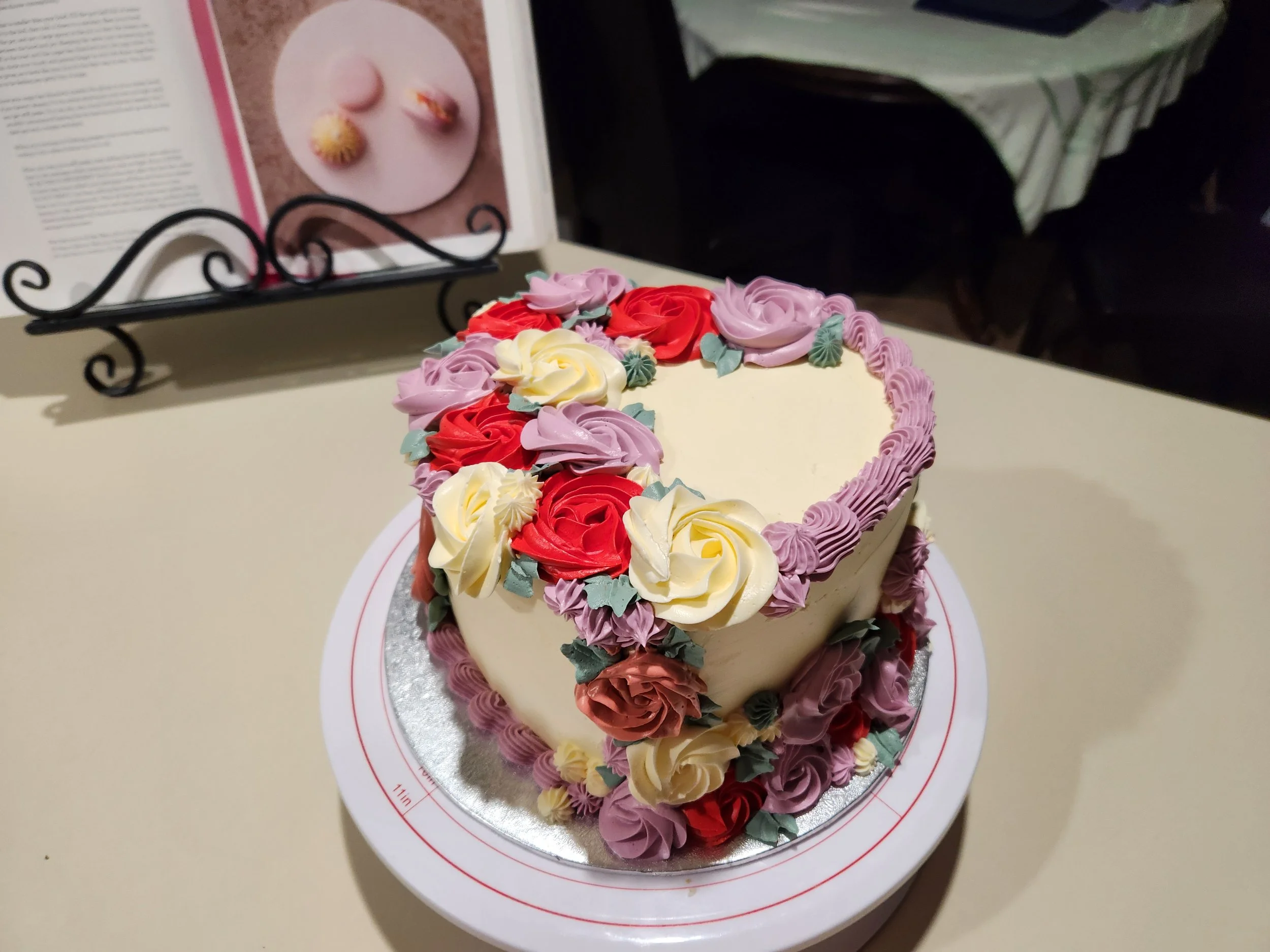Heart-shaped cake decorated with colorful icing roses in yellow, red, purple, and pink, with lavender piping around the border, on a white cake stand.