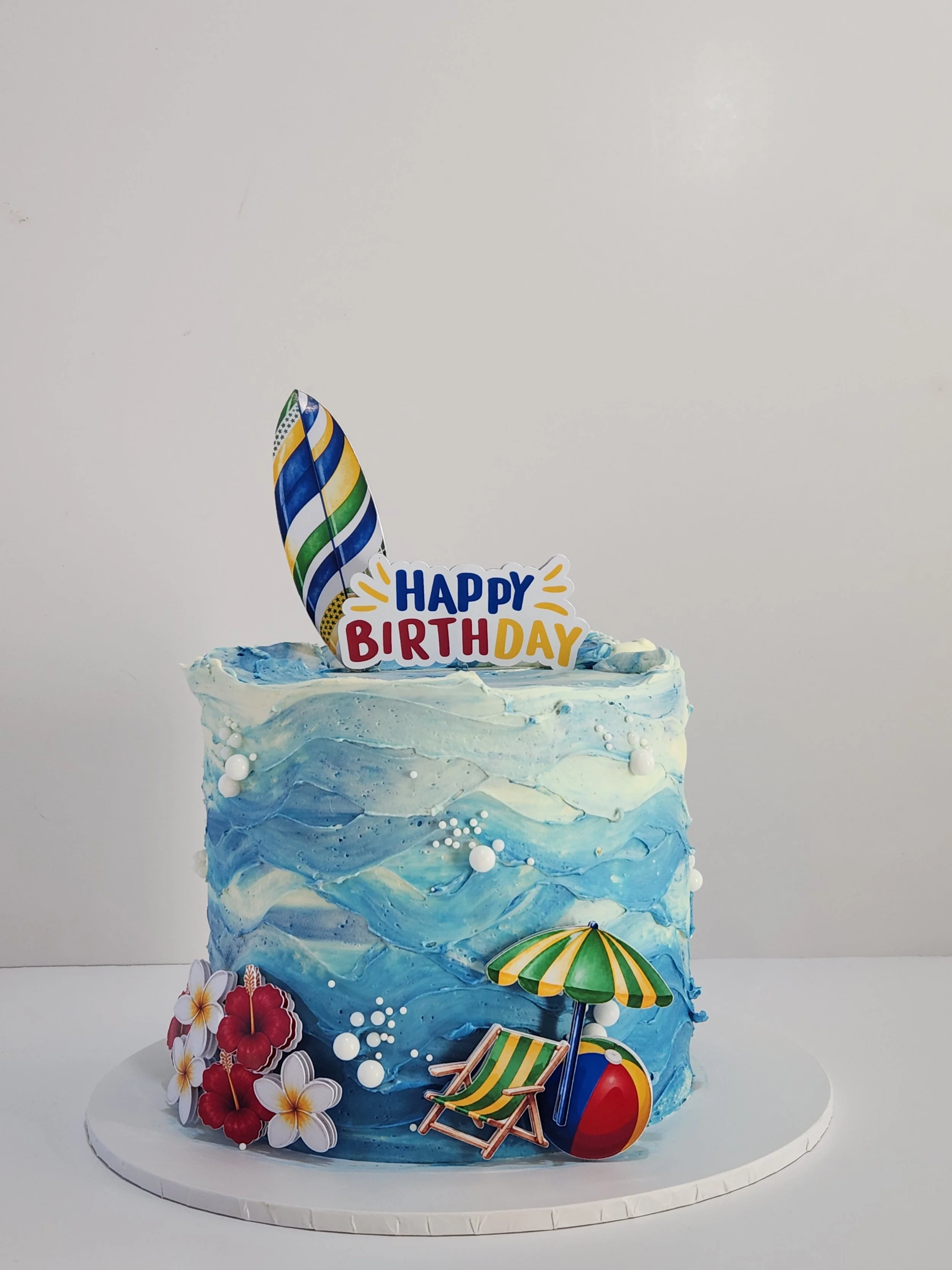 A multi-layered birthday cake decorated with ocean and beach themed items, including a colorful surfboard, umbrella, beach chair, and tropical flowers, with a 