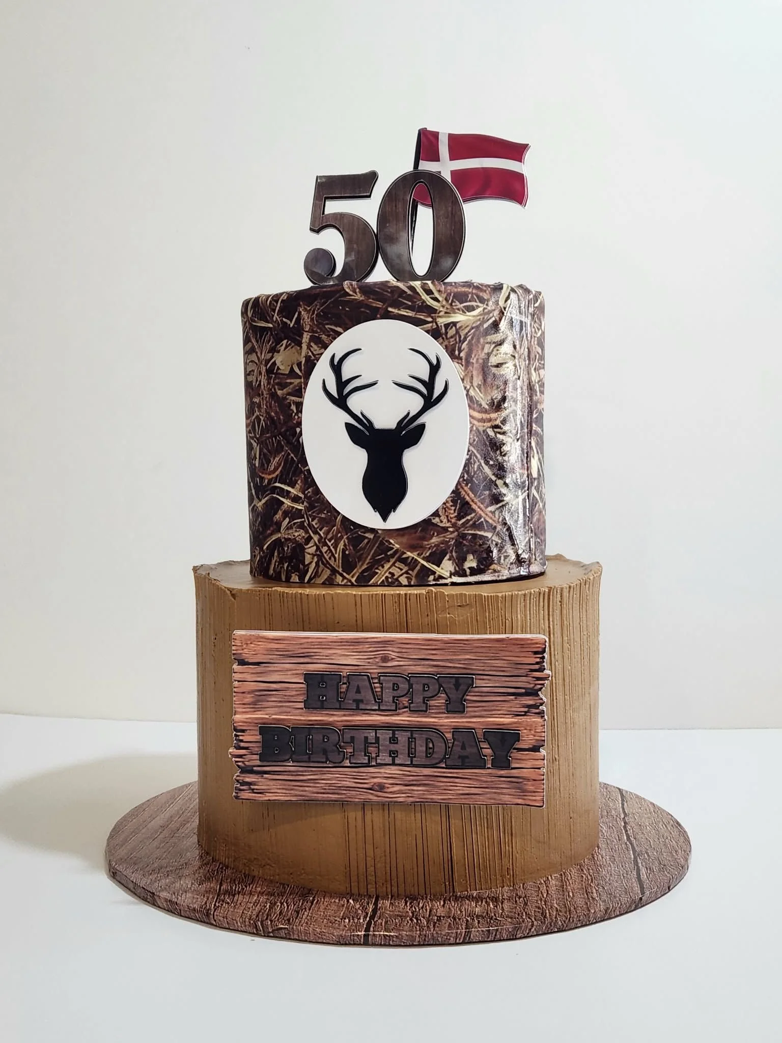 Birthday cake decorated with a camouflage pattern, a black silhouette of a deer head, and a '50' number topper with a Danish flag, placed on a wooden platform with a "HAPPY BIRTHDAY" sign.