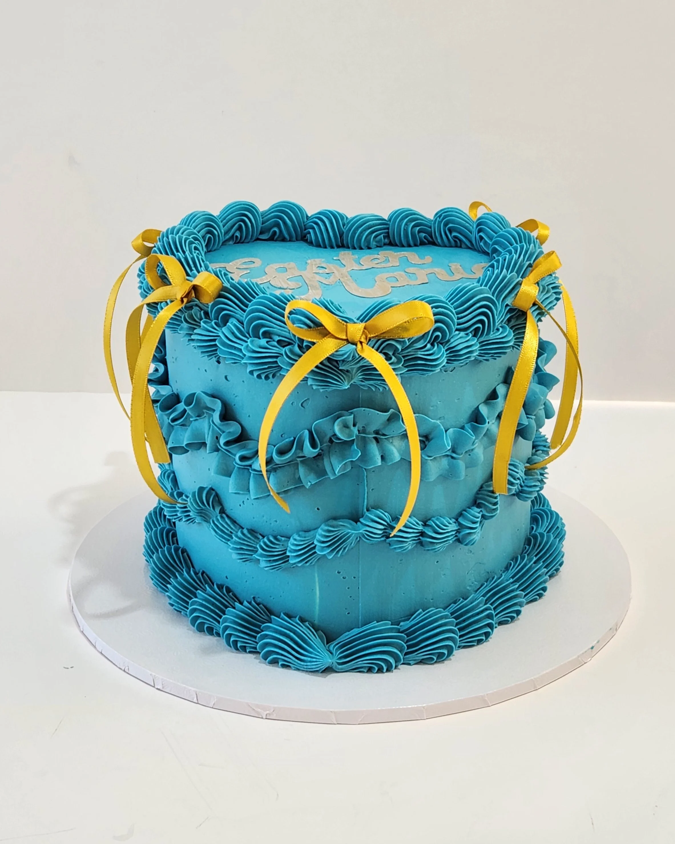 Blue birthday cake decorated with piped blue frosting and yellow ribbons. The top of the cake has the words "Happy Birthday" written in white.