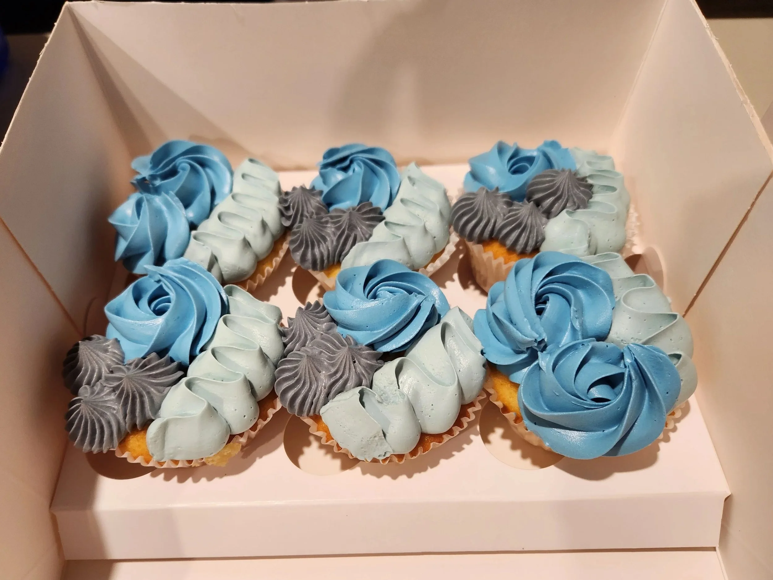 A white box containing twelve cupcakes with pastel-colored frosting, some with blue and some with grayish-green swirls, arranged in three rows.