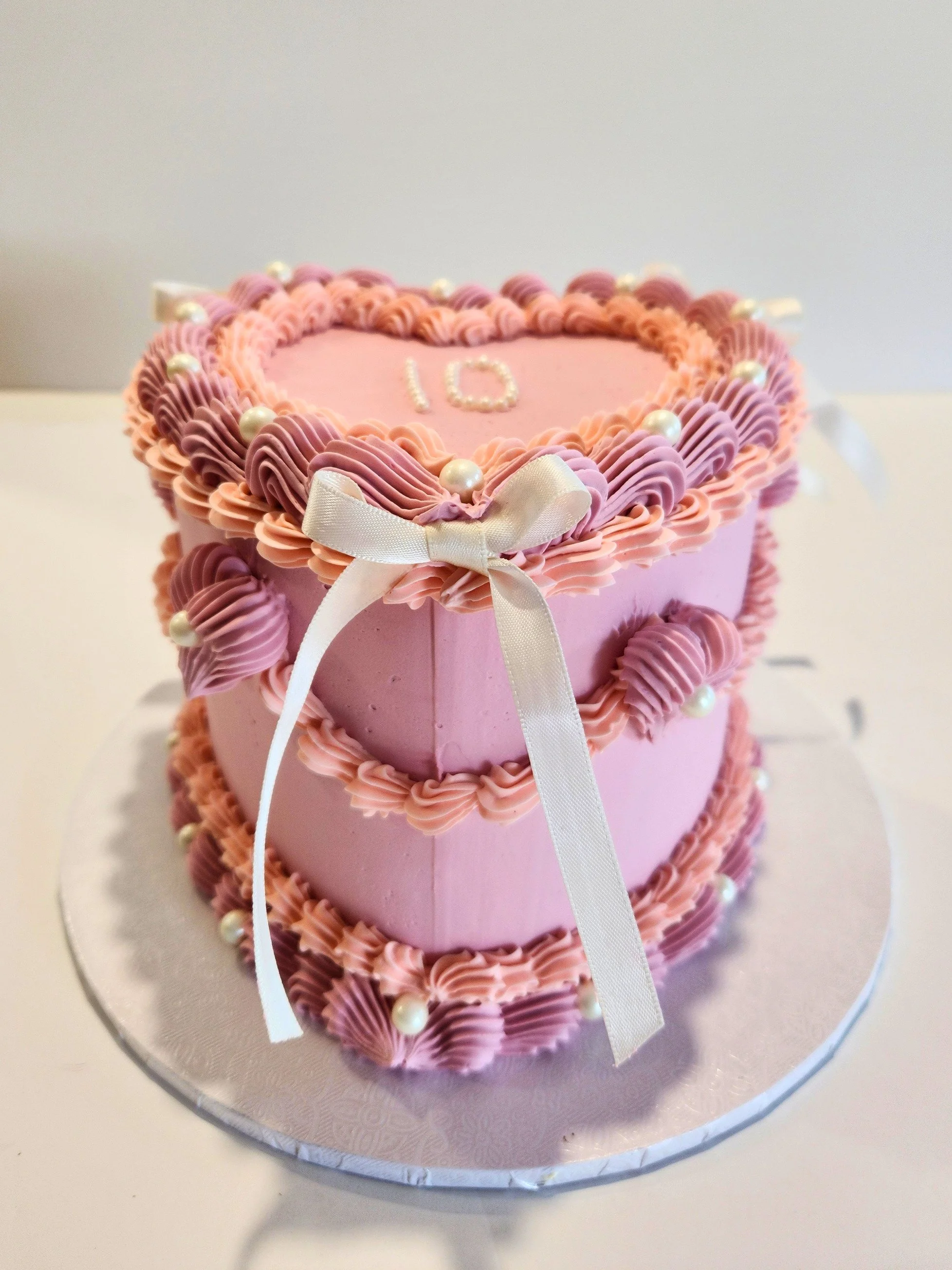 Pink heart-shaped cake decorated with two tone pink frosting, white pearls, and a white ribbon bow, with the number 10 on top.
