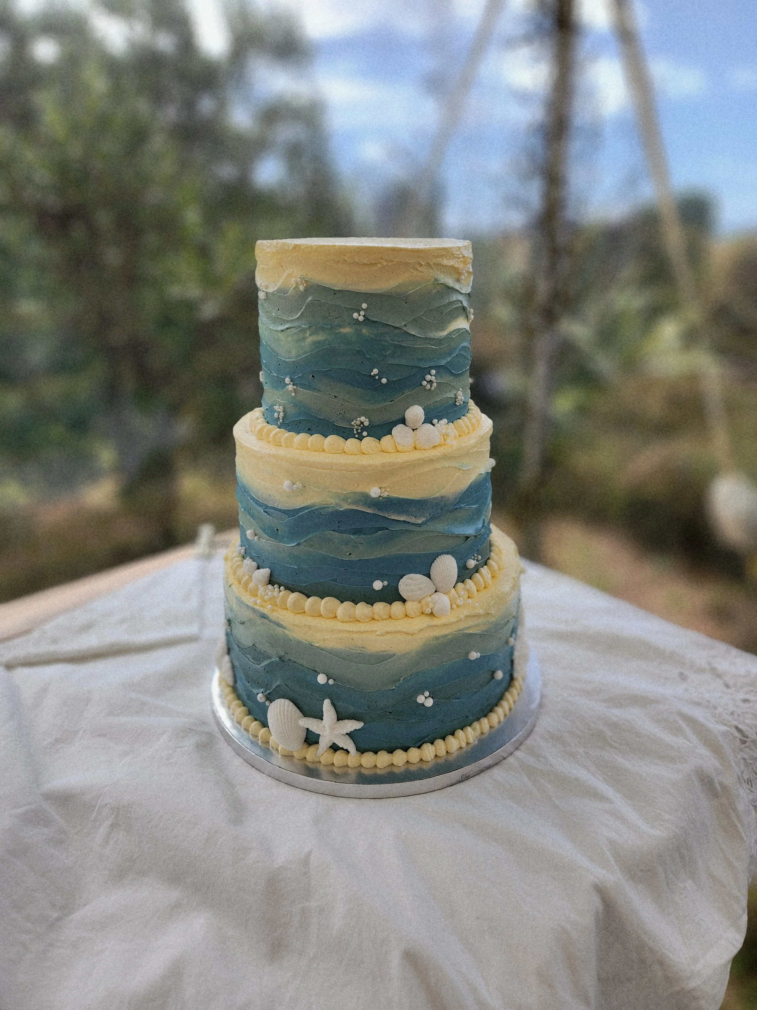 Planning a beach wedding requires a balance of natural beauty and refined detail. Nothing anchors a seaside reception quite like a custom-designed three-tiered cake that mirrors the rhythm of the Pacific Ocean.