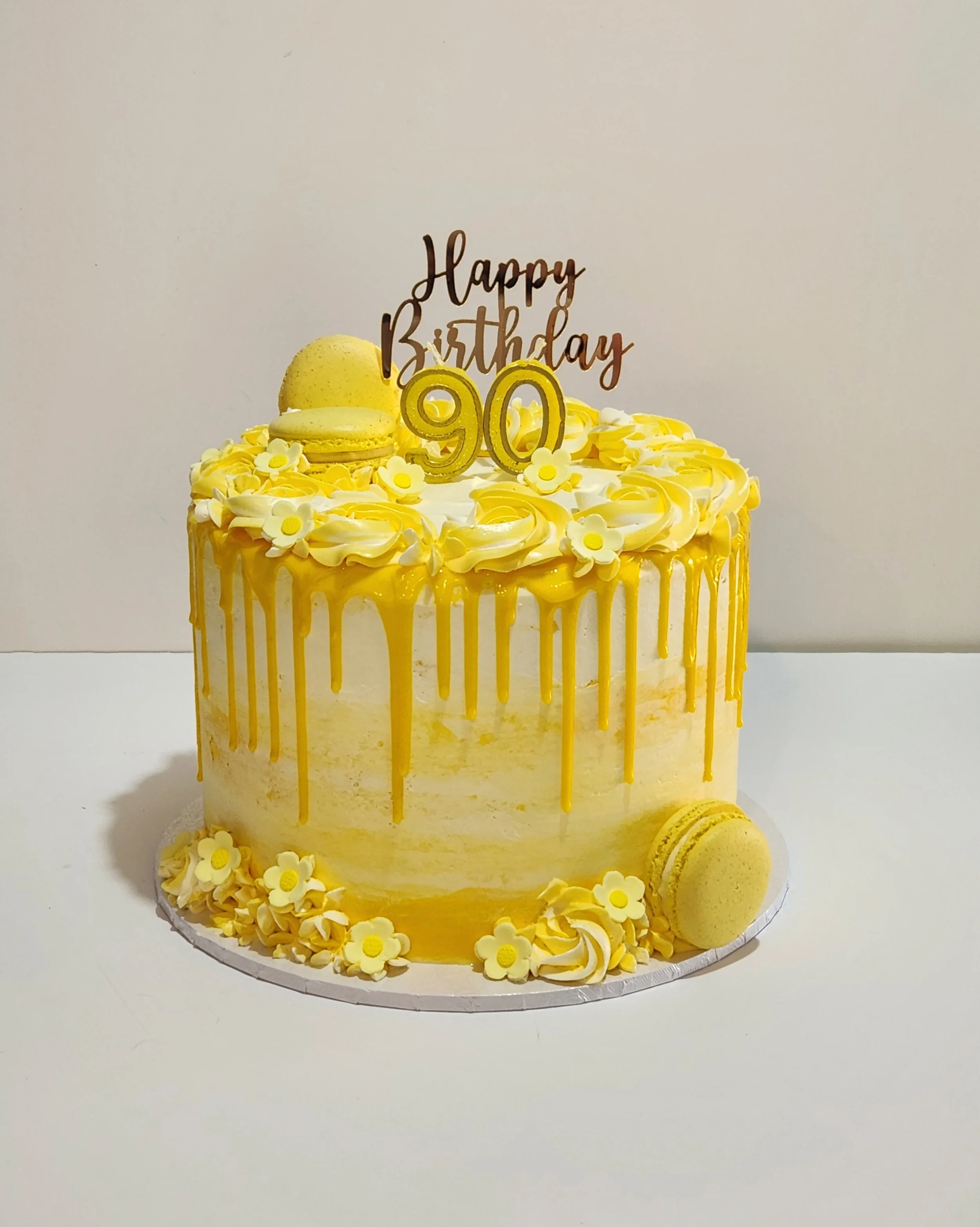 Yellow birthday cake with white and yellow floral decorations, macarons, and a "Happy Birthday" topper reading 90.