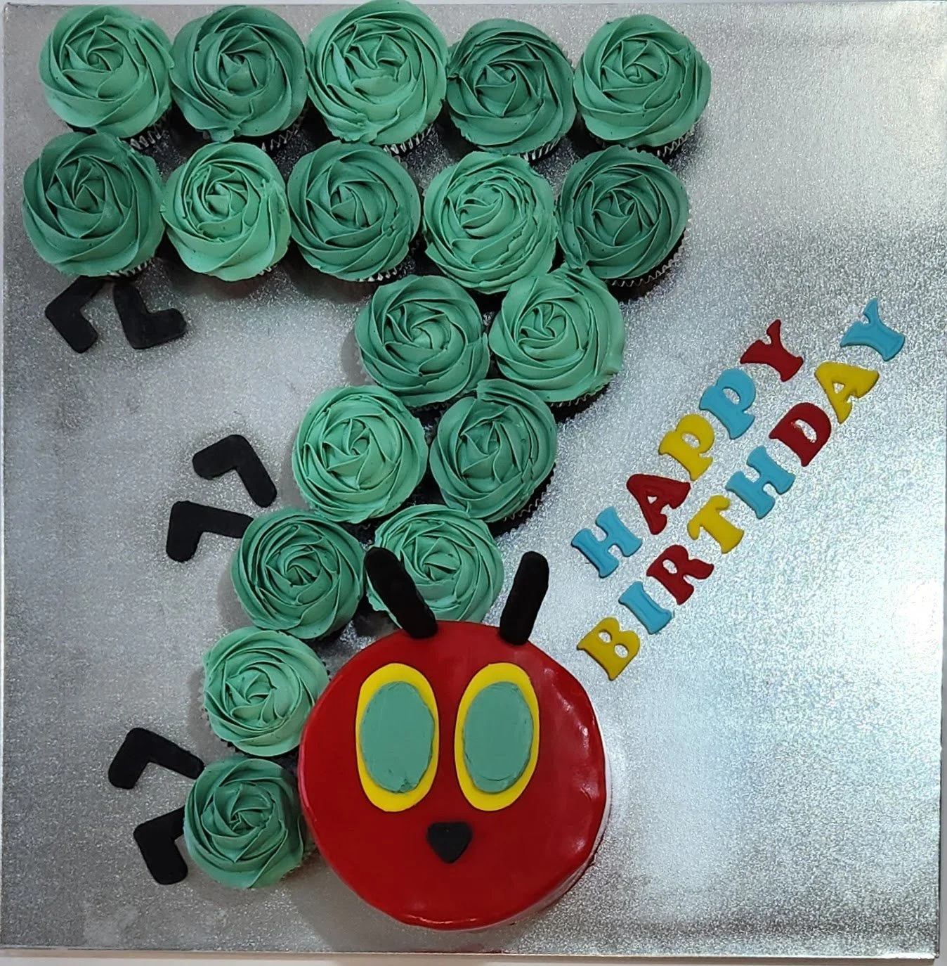 Cake decorated for a birthday celebration with a figure of a red character with yellow and blue eyes, black antennae, and a small black nose. Green frosting roses form the body, with the number seven written in black icing. The words 'Happy Birthday'