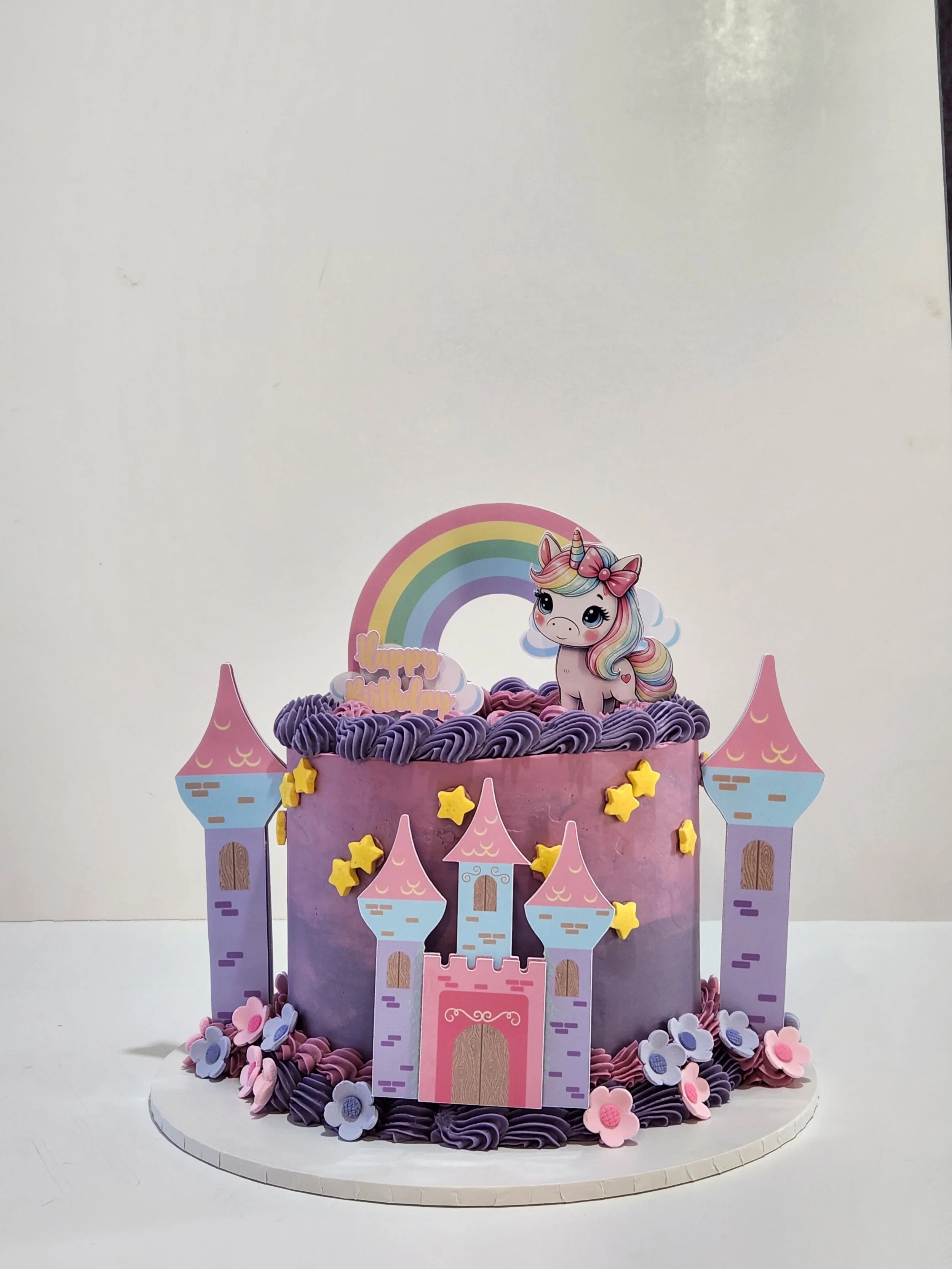 Colorful unicorn-themed birthday cake with a castle design, purple and pink frosting, and a unicorn figurine on top, surrounded by flowers.