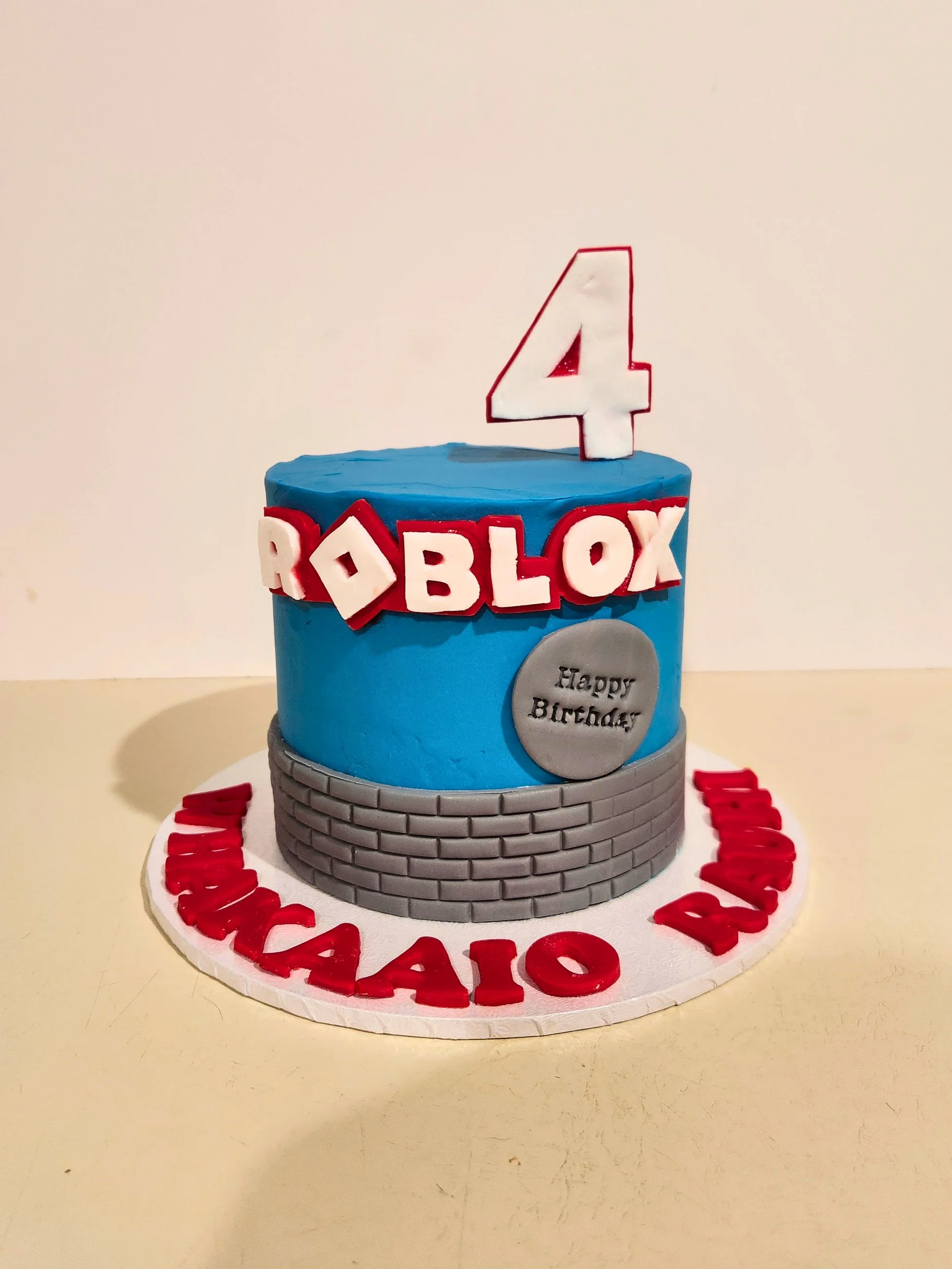 A roblox birthday cake with blue icing, red and white decorations, a large number 4 candle, and a 'Happy Birthday' message on a gray coin-shaped topper. The cake is on a white round base with red lettering.