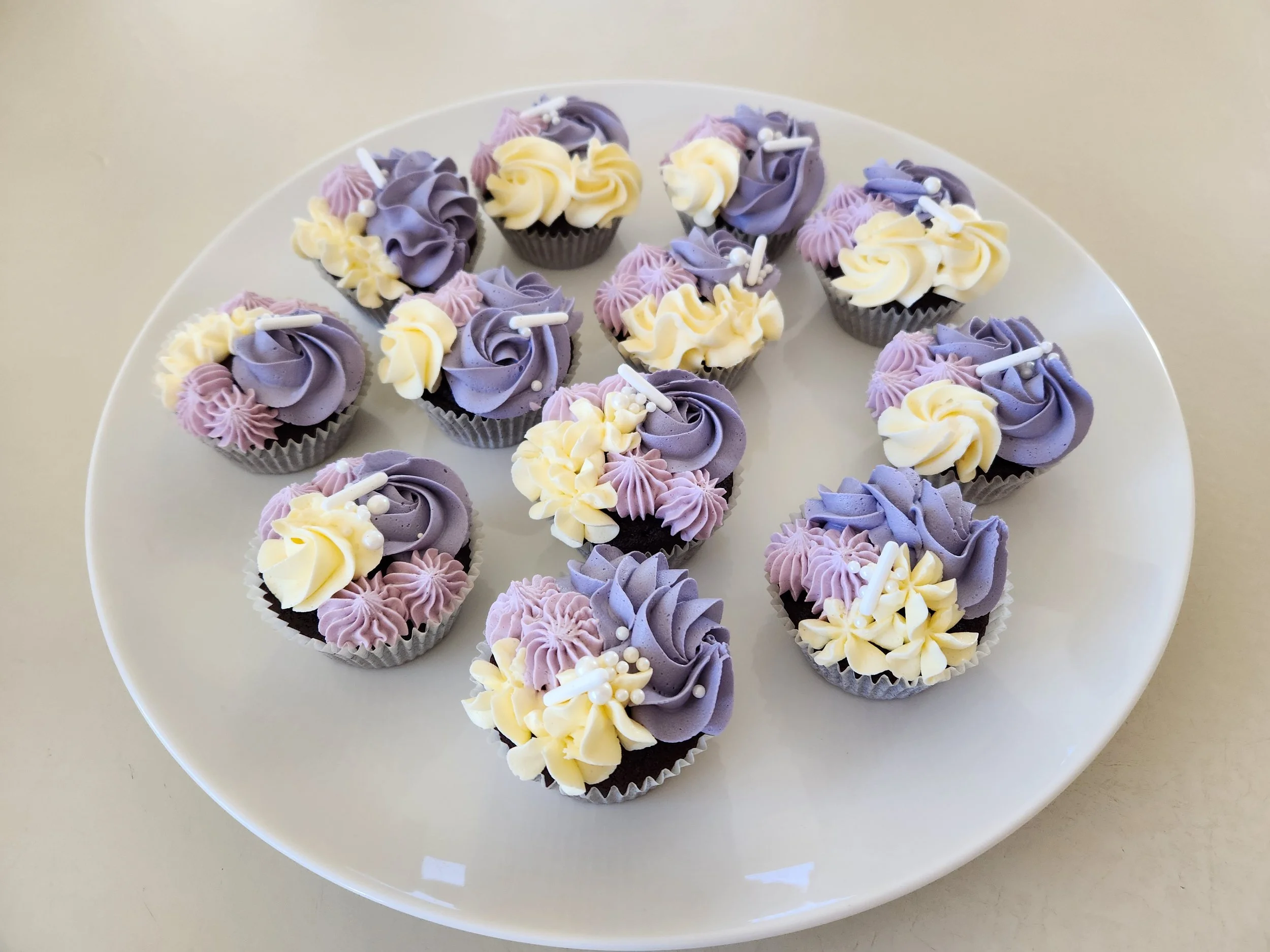 Plate of assorted purple, white, and light pink cupcakes decorated with swirls of frosting and small edible pearls and sticks.