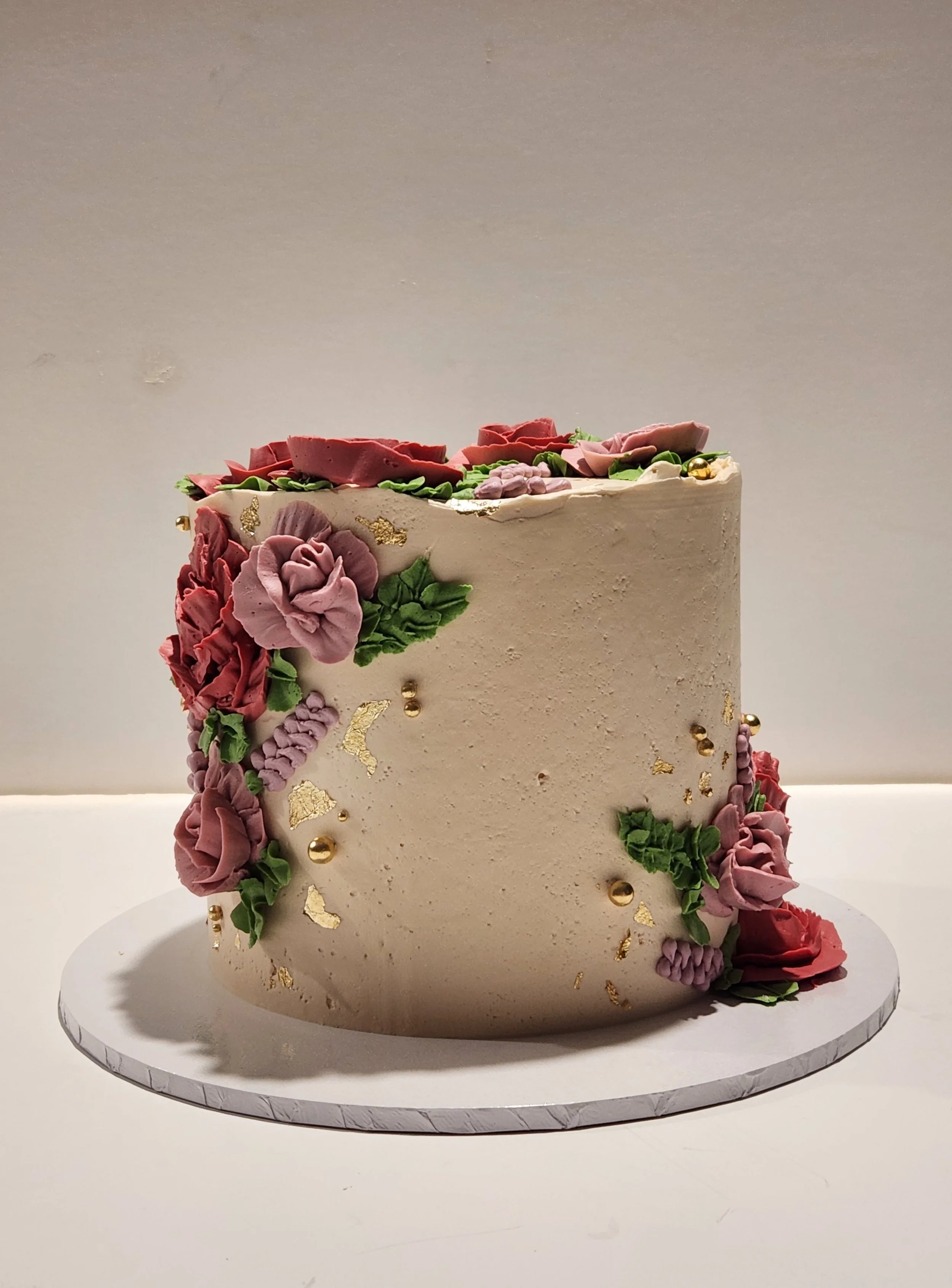 A tall, white cake decorated with pink, purple, and green floral frosting and gold accents, placed on a white marble cake board.