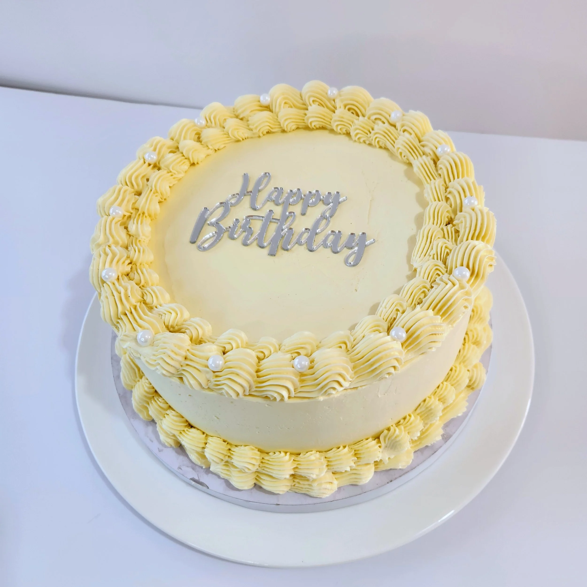 Yellow birthday cake with piped frosting border and pearl decorations, with the words 'Happy Birthday' written in silver on top.