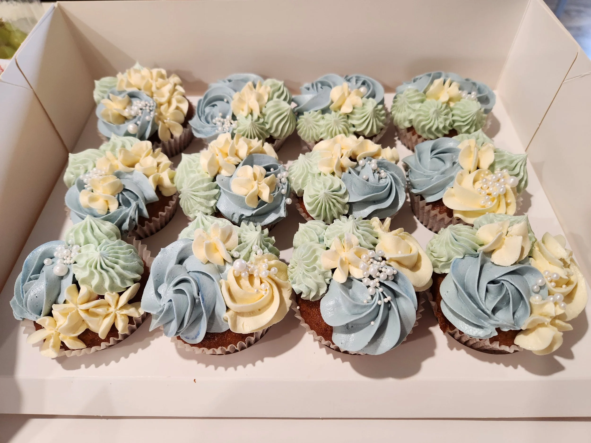 A box of designer cupcakes with pastel blue, green, and yellow swirled icing topped with edible pearls and small flower decorations. Vanilla with Berry compote filling and Vanilla Swiss Meringue Buttercream.