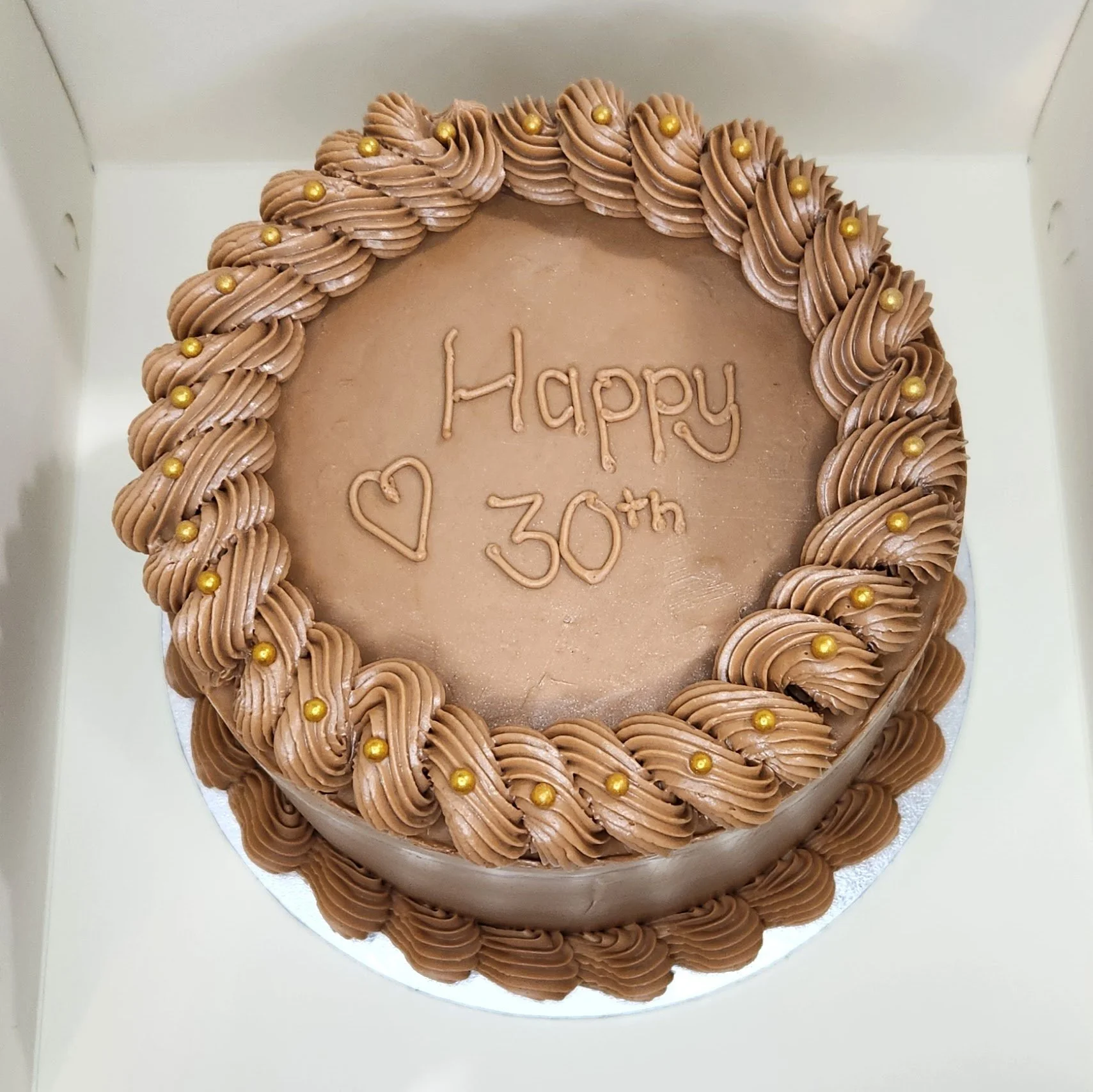 A round chocolate cake with chocolate frosting, decorated with piped rosettes and small gold beads around the edges, with the message "Happy 30th" written in icing and a heart drawn underneath.