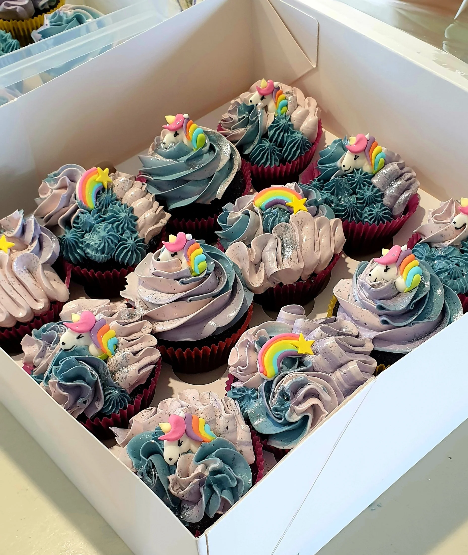 Decorated cupcakes with unicorn and rainbow toppers in a white box.