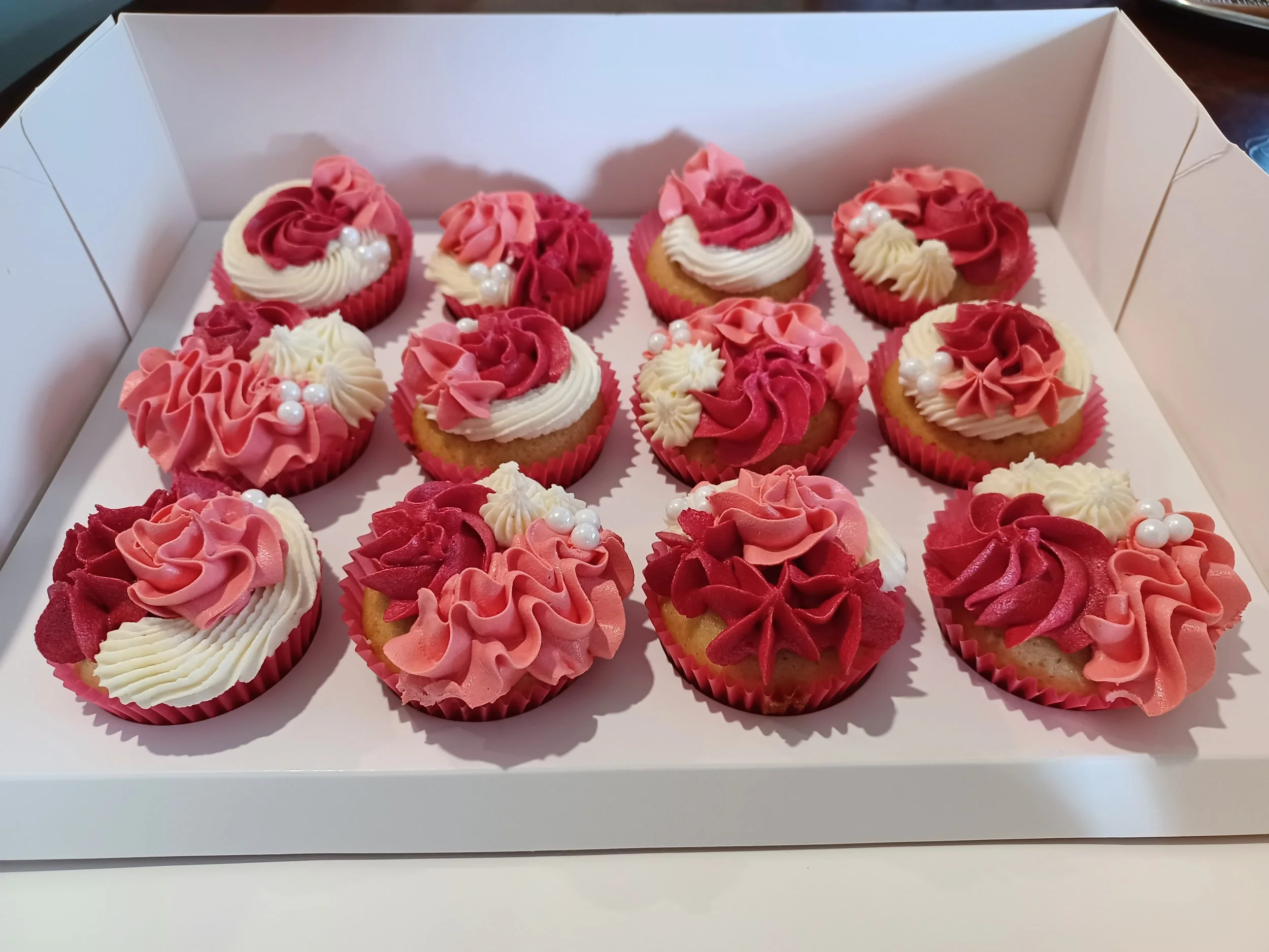 A white box containing twelve decorated cupcakes with pink, red, and white frosting and pearl-like sprinkles.
