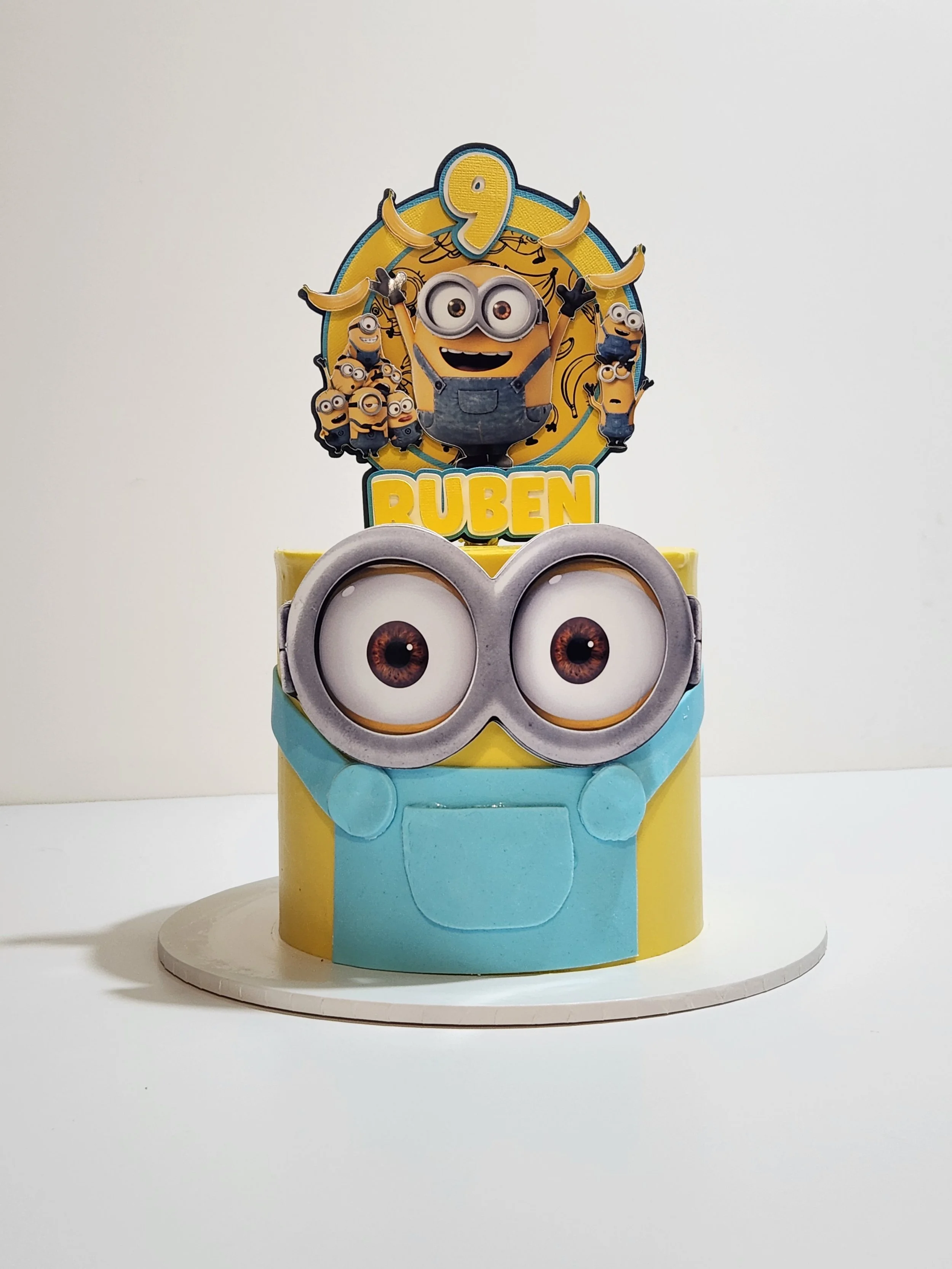 Minion themed birthday cake with Minion figures and a blue and yellow color scheme, topped with a banner reading '9' and the name 'Ruben'.