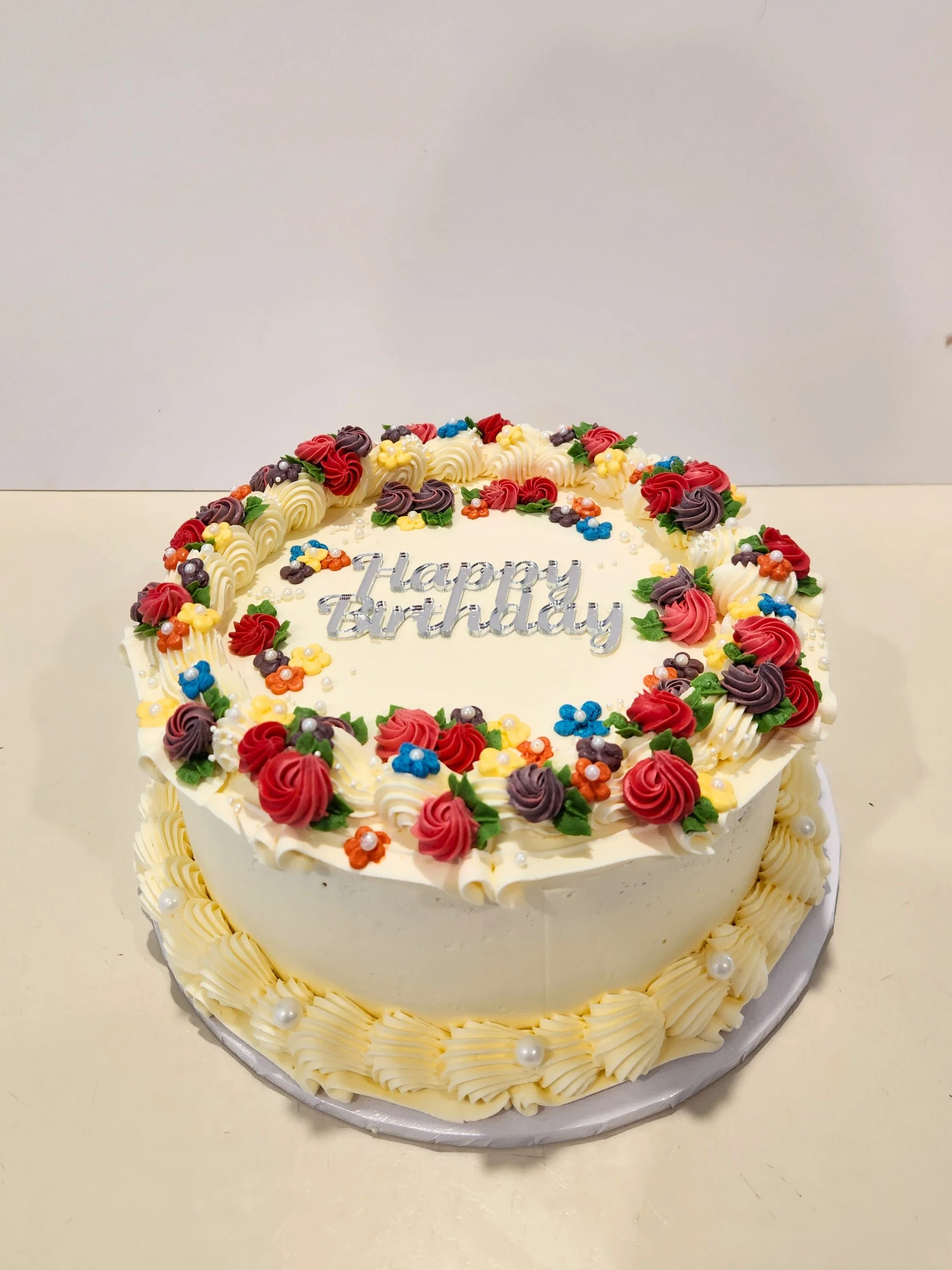 Round birthday cake decorated with multicolored flower-shaped icing, green leaves, and pearl-like spheres, with a silver "Happy Birthday" topper.