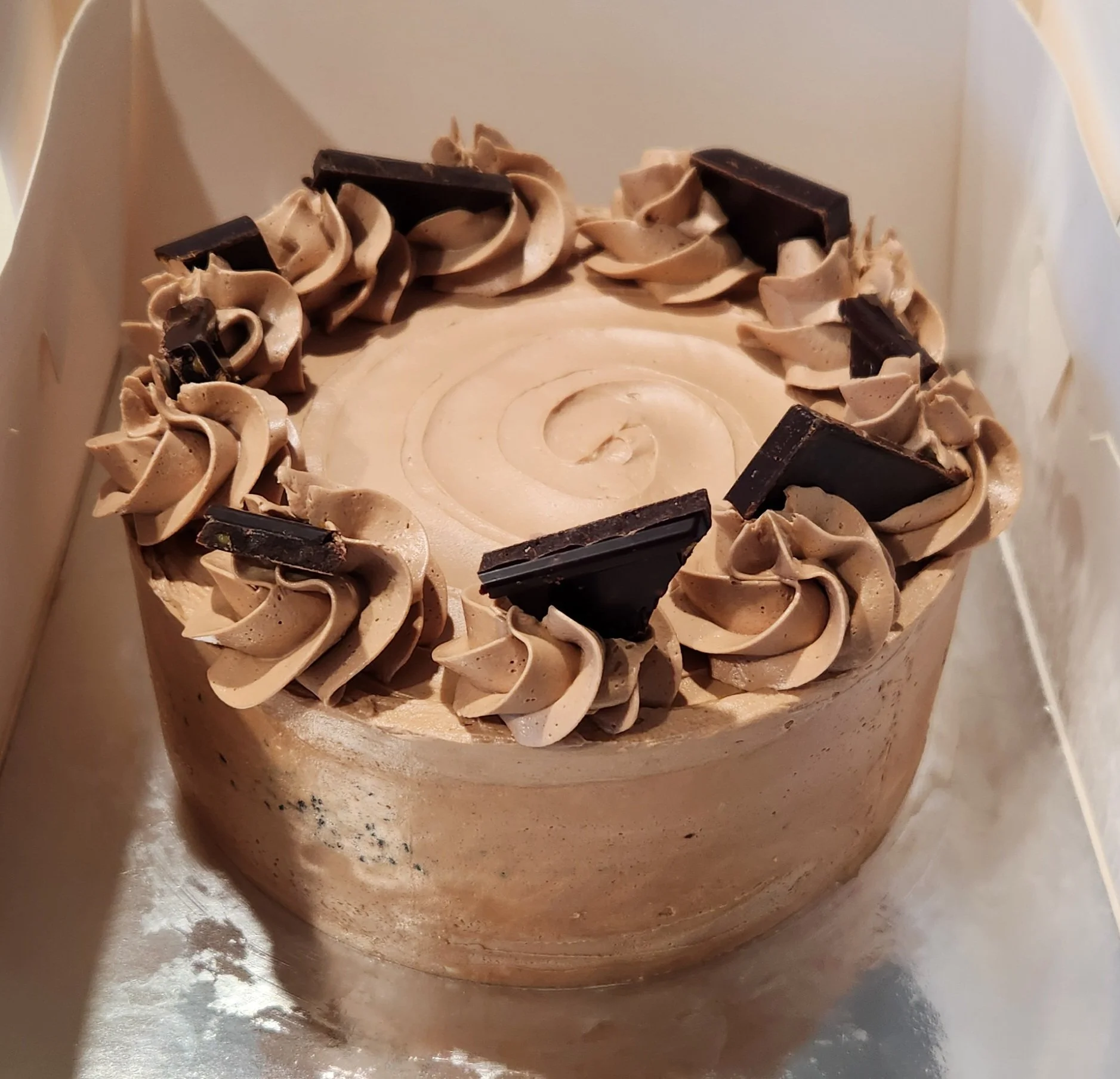 Close-up of a cake decorated with swirls of beige and brown frosting, with dark chocolate pieces around the top edge.