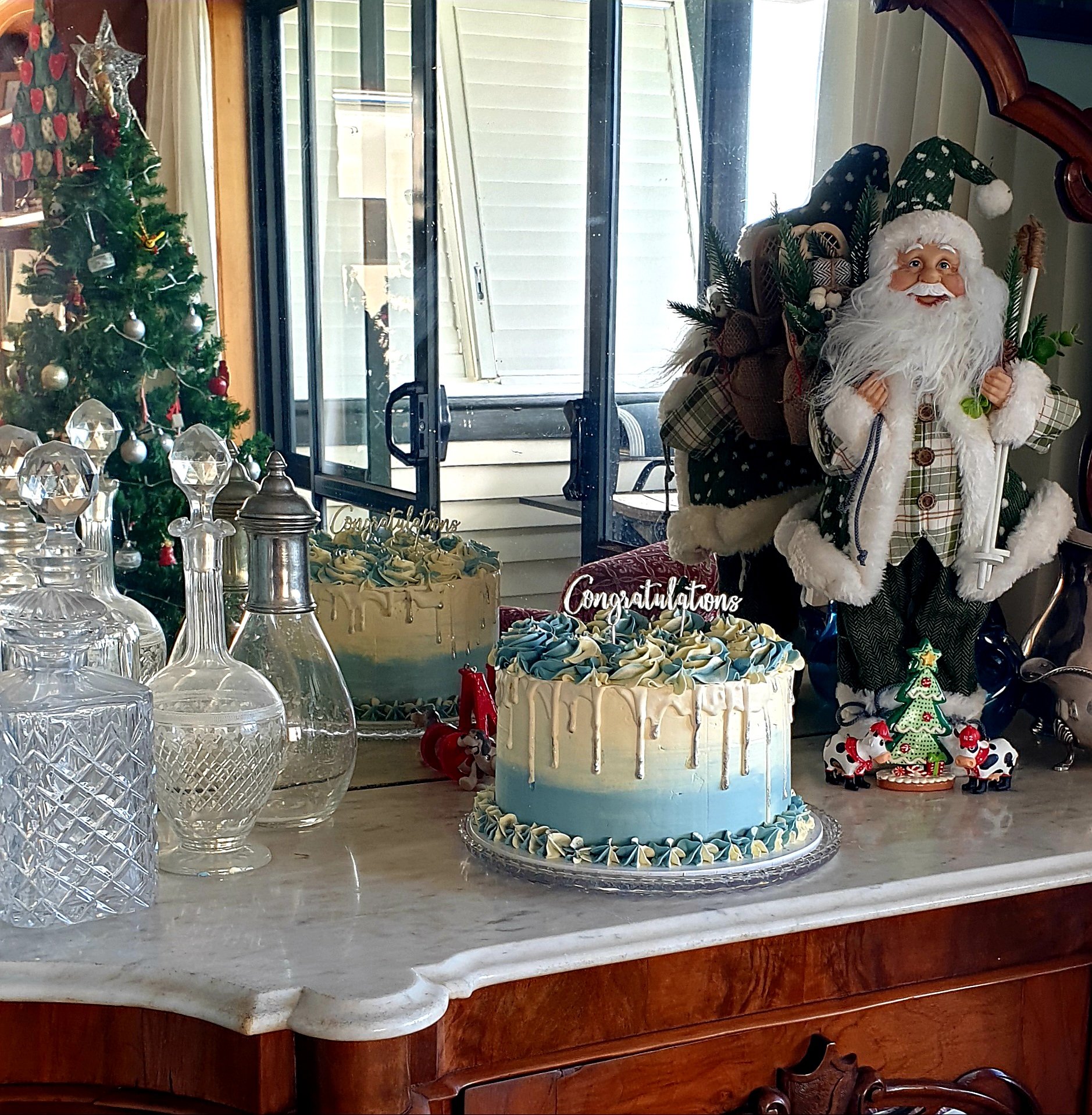 A Christmas-themed display on a wooden table, featuring a decorated Christmas tree on the left, several glass and metal decorative items, and two cakes, one with white and blue frosting. There is a Santa Claus figurine on the right, dressed in plaid 