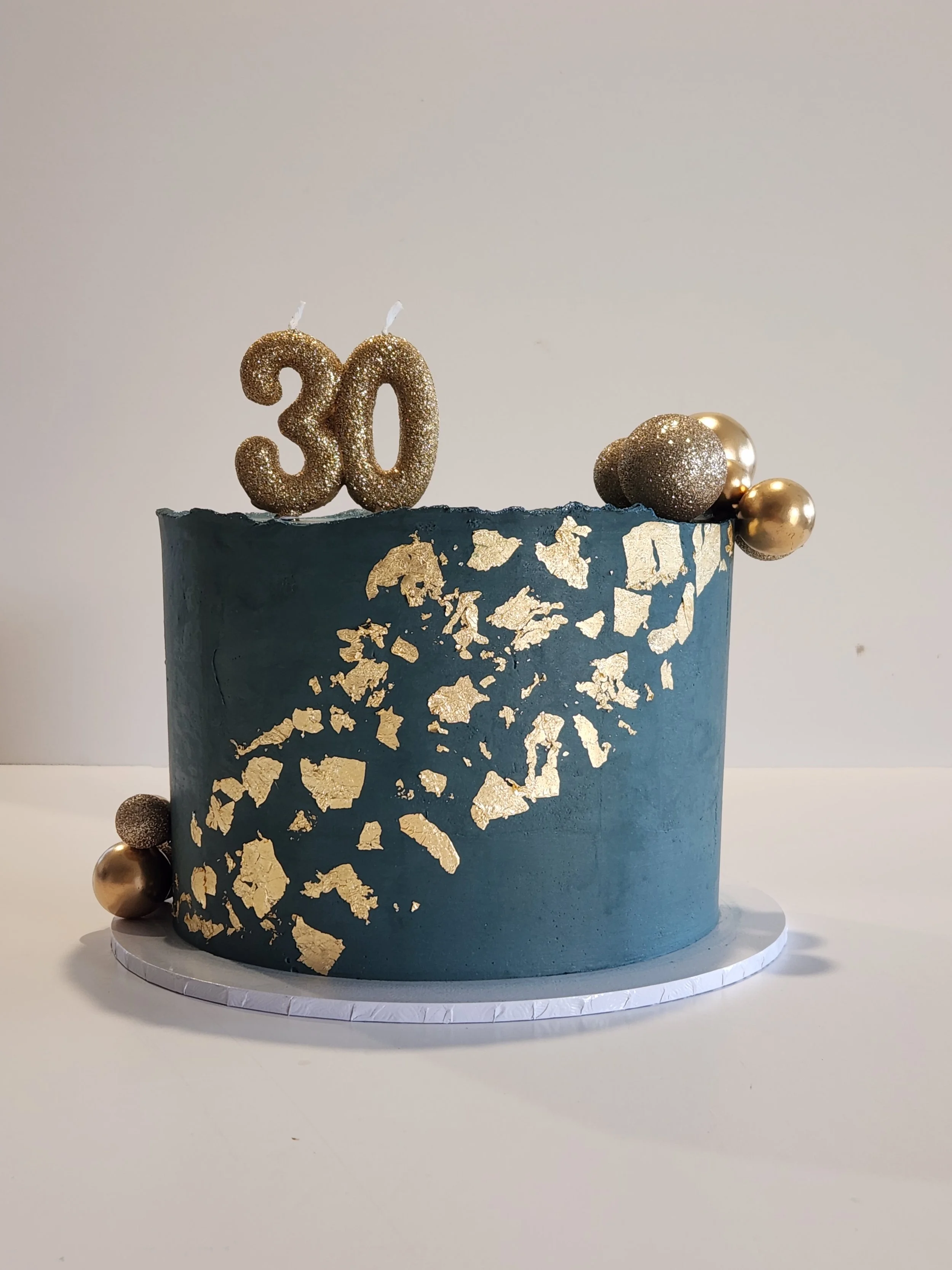 A round blue cake decorated with gold leaf accents, gold and glittery gold number candles forming the number 30, and gold and glittery gold spherical decorations on top.