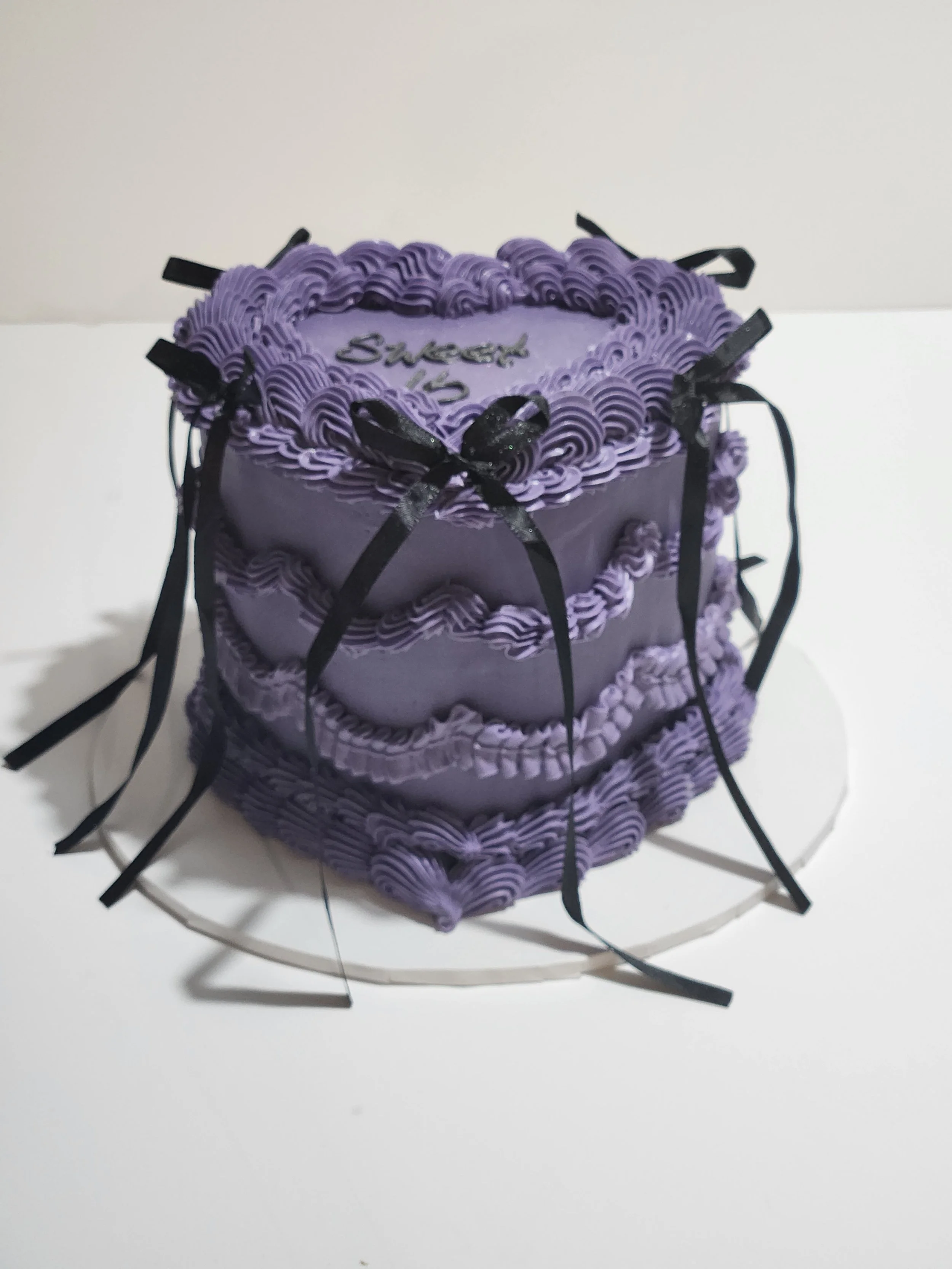 A purple birthday cake decorated with purple frosting and black ribbons, with a message written on it, placed on a white wire cake stand against a plain light background.