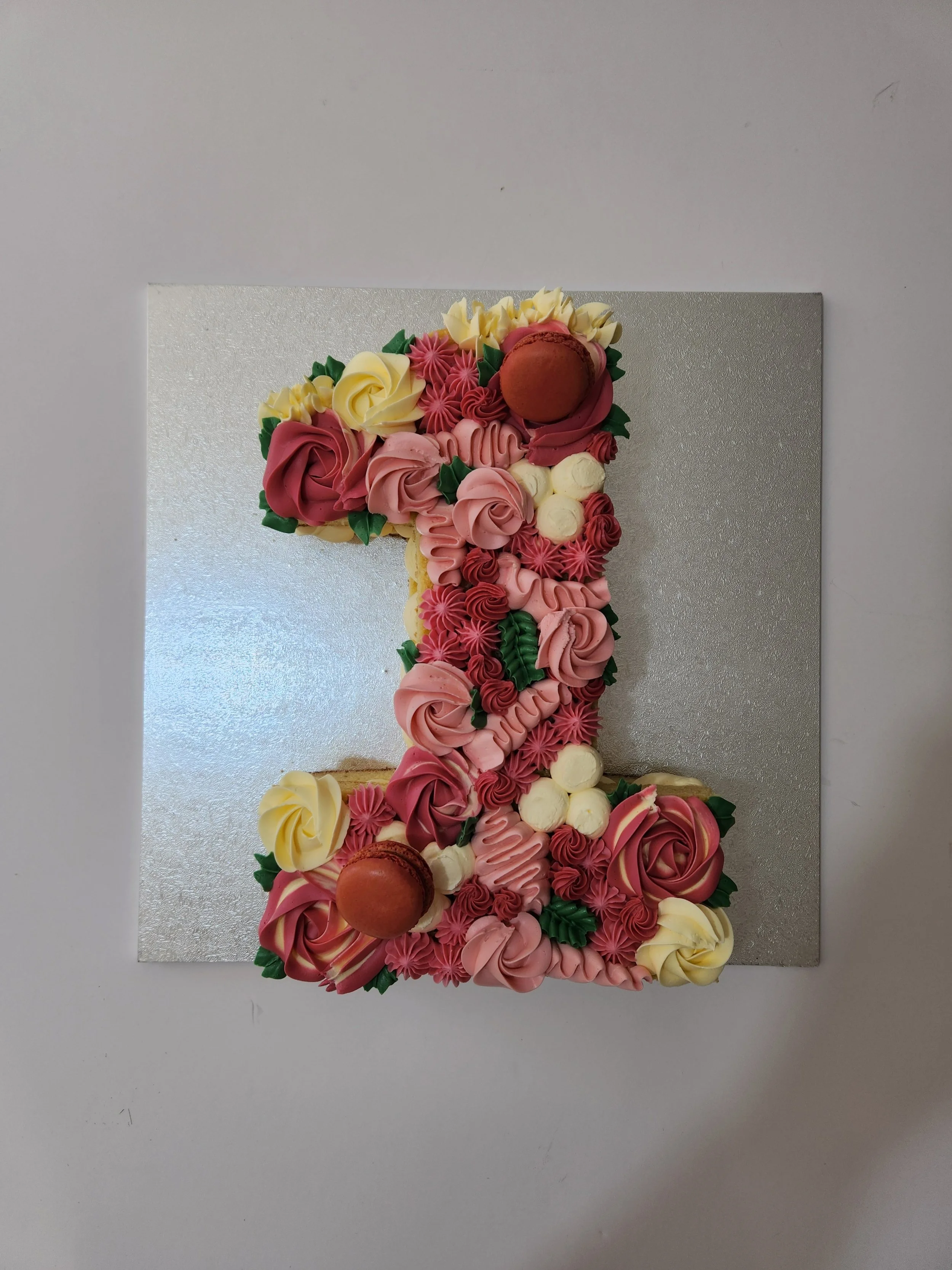 Decorative number 1 cake with pink, yellow, and red frosting roses, piped flowers, a chocolate macaron, and green leaves, on a silver board.