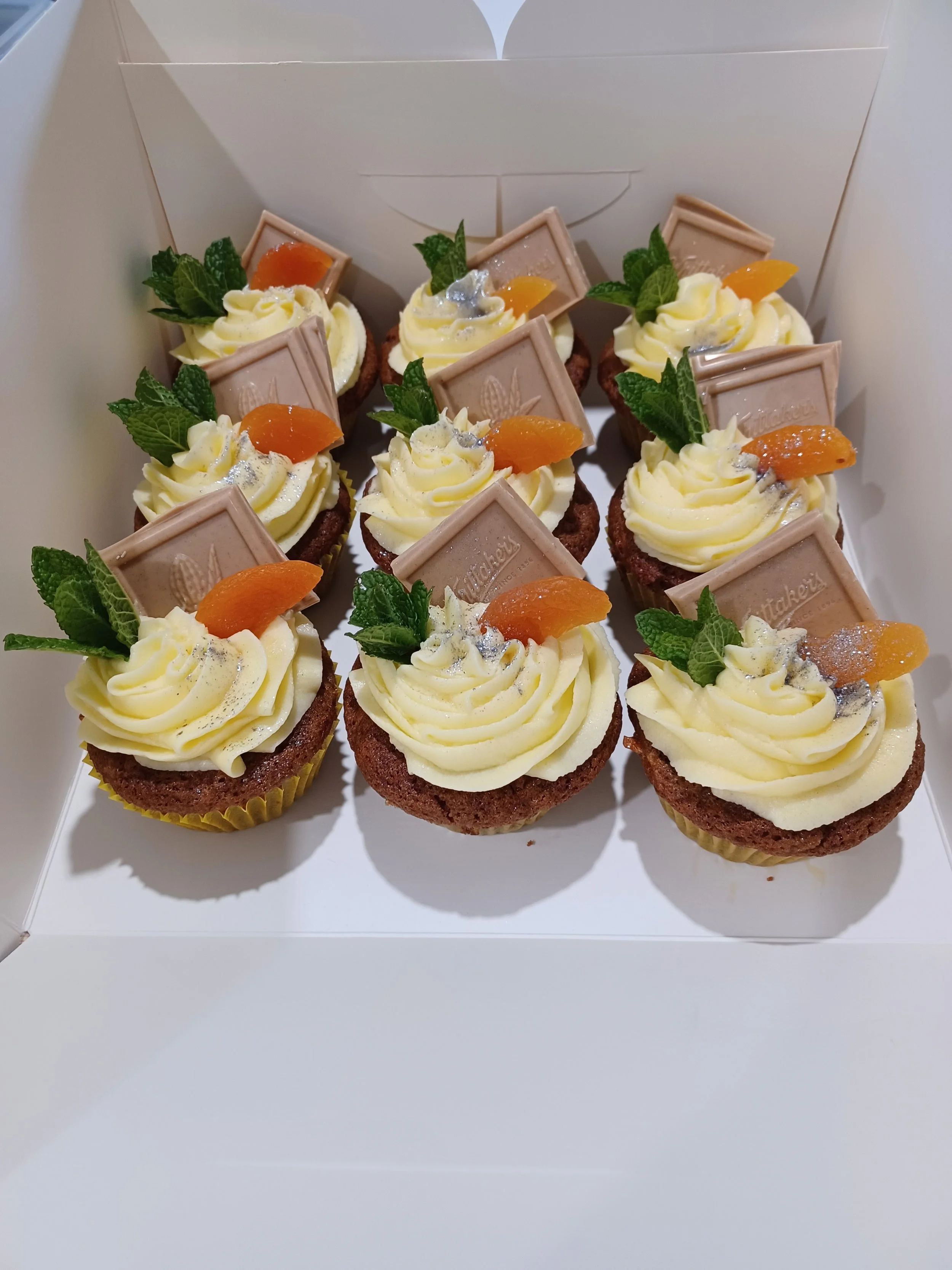 Six cupcakes topped with yellow frosting, garnished with a small piece of orange fruit, fresh mint leaves, and a small piece of chocolate.