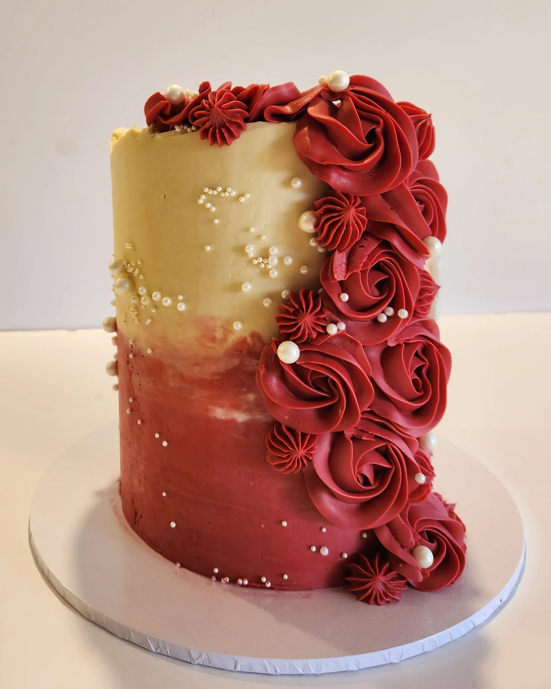 A tall, cylindrical birthday cake decorated with red and white accents. The cake features red buttercream roses and piped decorations along the side, with white pearls and small edible beads adorning the surface.