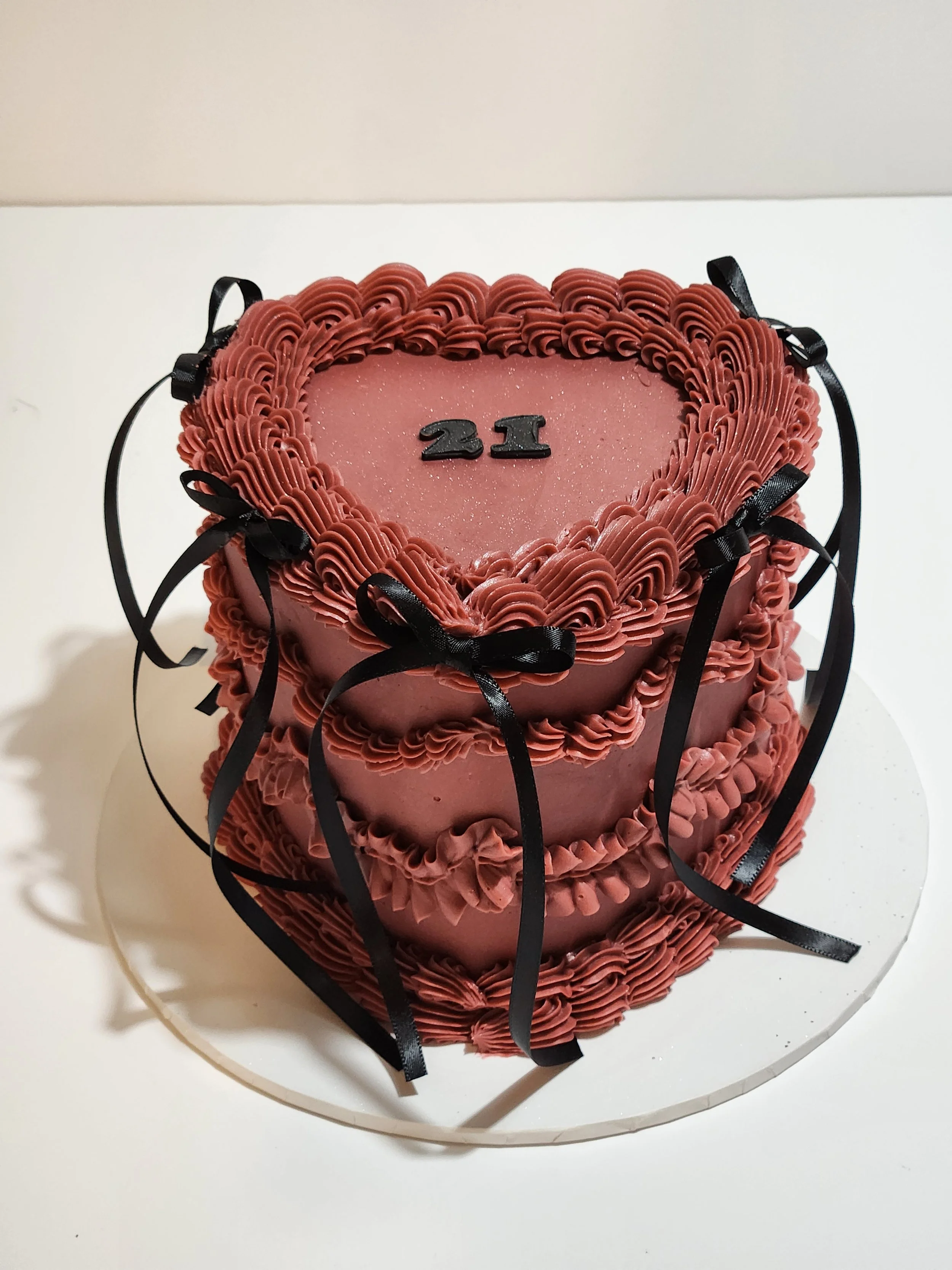 A pink, heart-shaped cake decorated with piped pink frosting and black ribbons. The top of the cake has the number '21' in black decorations.