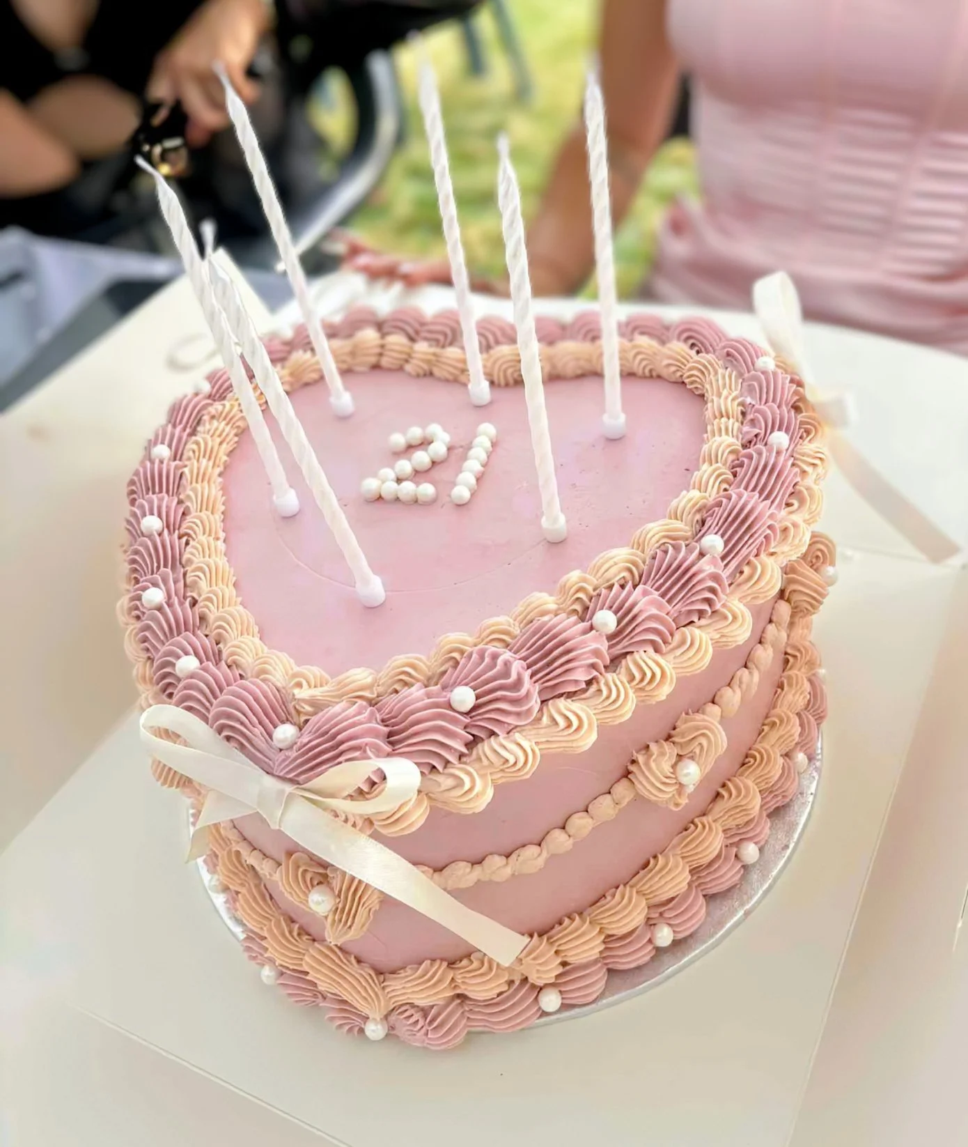 A pink, heart-shaped birthday cake decorated with light pink and darker pink frosting, white pearl-like candies, and pink and white candles. The number 21 is spelled out with small white candies on top.