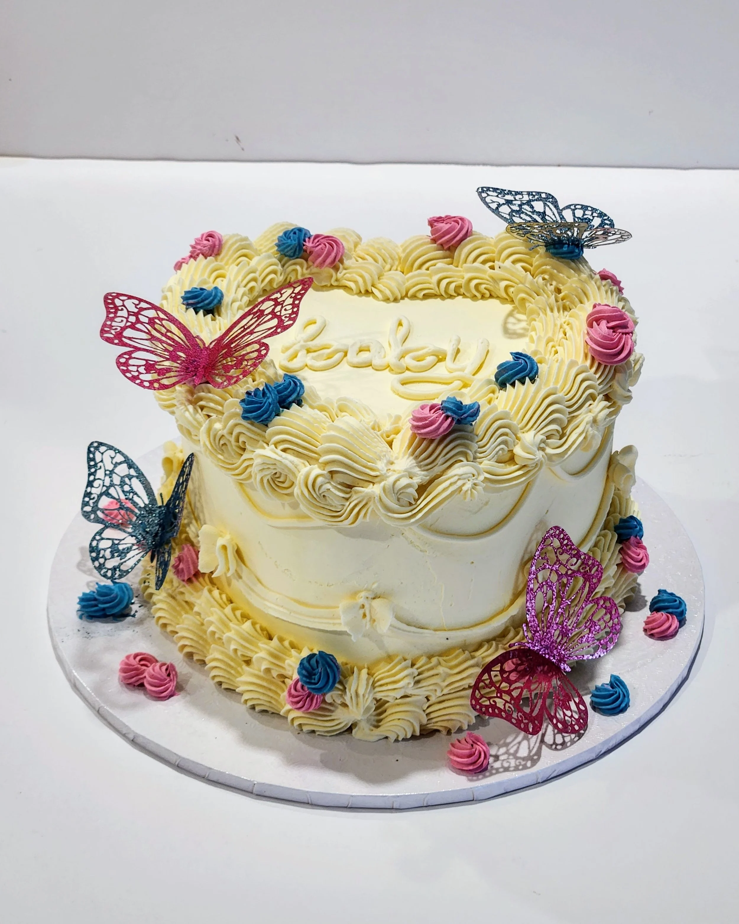 Vintage heart cake with off-white buttercream, pink and blue flower decorations, and pink and blue butterfly cutouts on top and around the cake, gender-reveal cake.