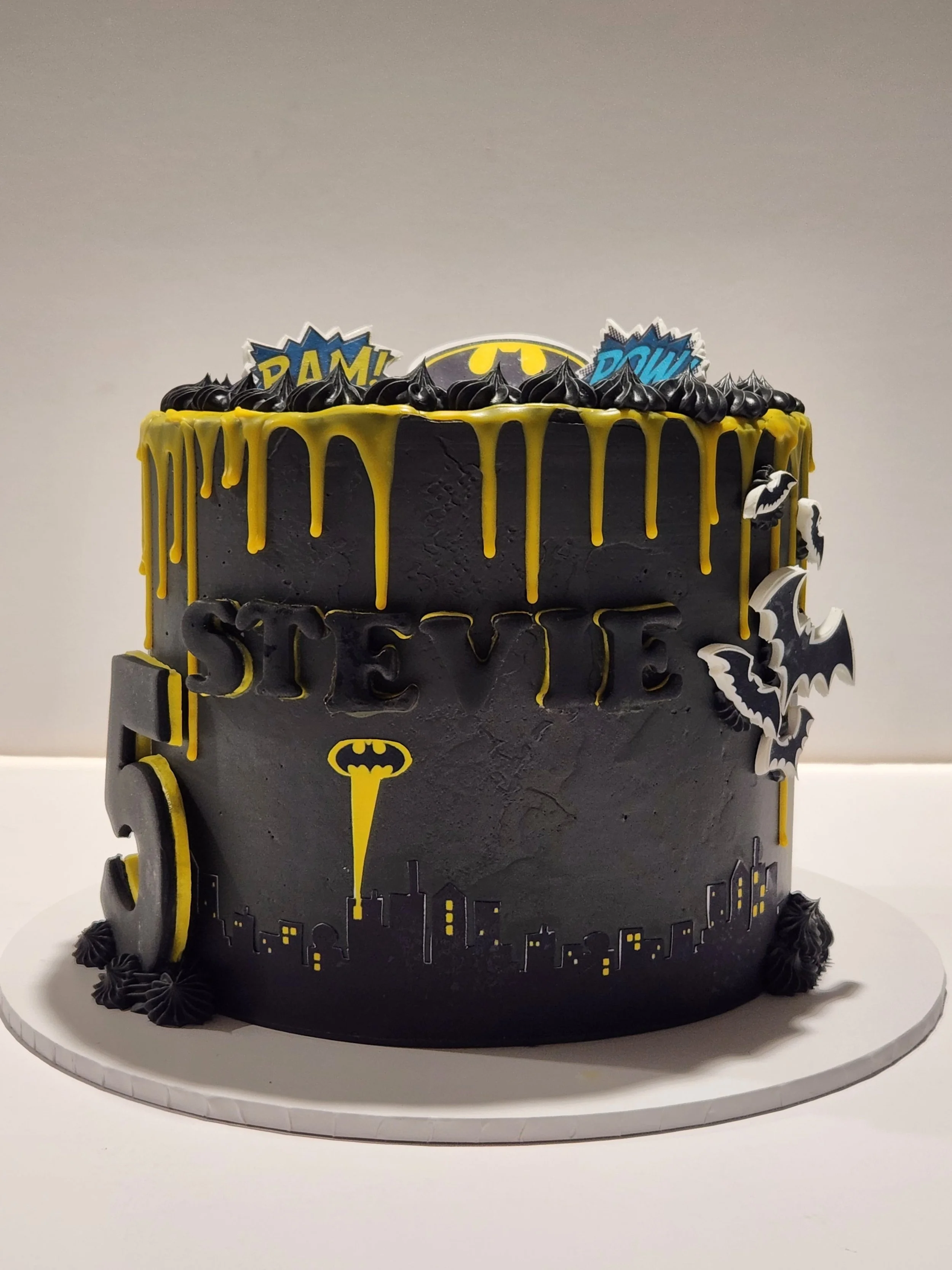 A Batman-themed birthday cake decorated with black and yellow colors, featuring Batman logos, cityscape and comic book style words like 'BAM!' and 'POW!'.