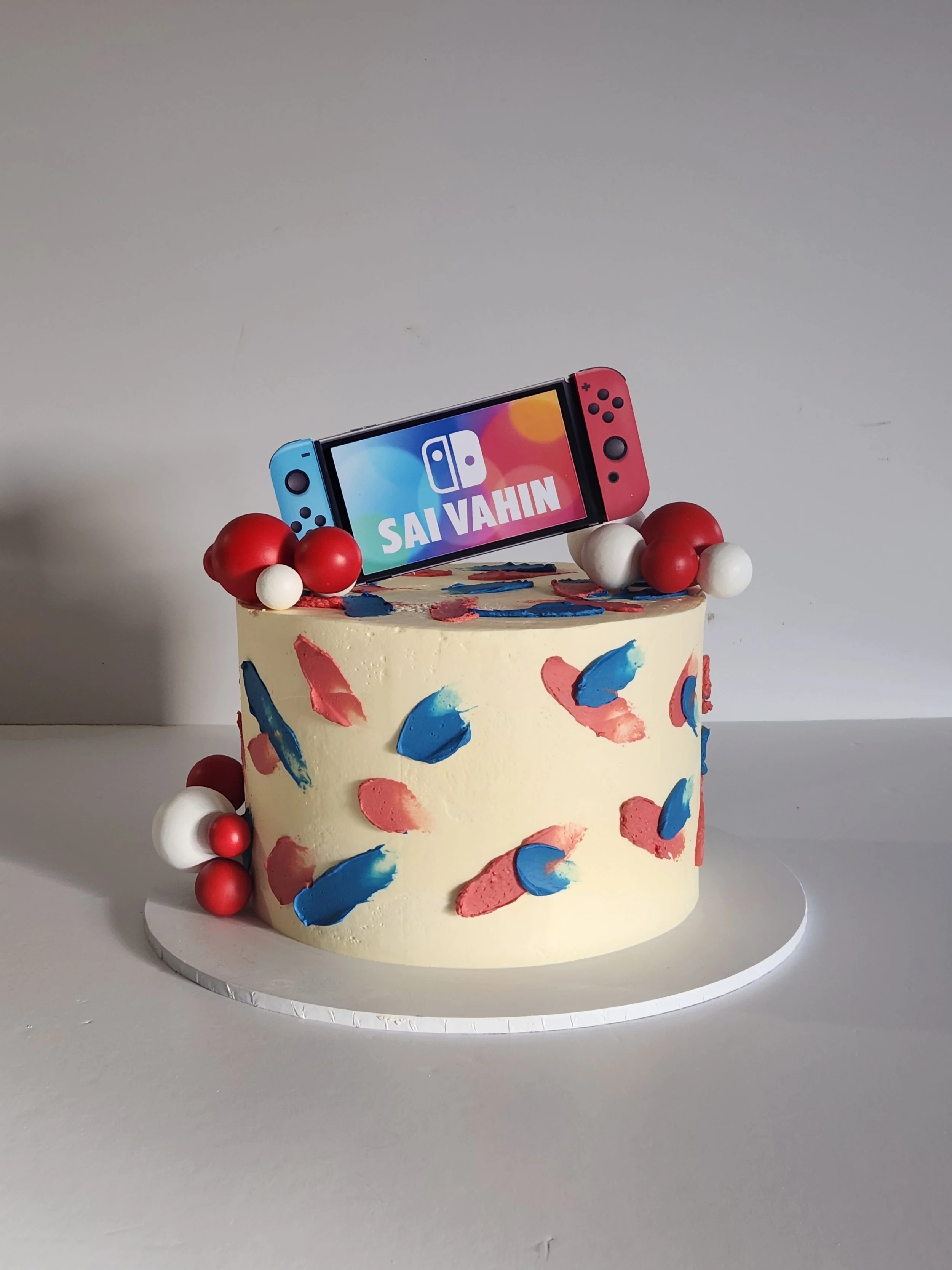 Decorative cake with colorful abstract paint strokes, red and white spherical decorations, topped with a Nintendo Switch console displaying the name 'Sai Vahin' on the screen.