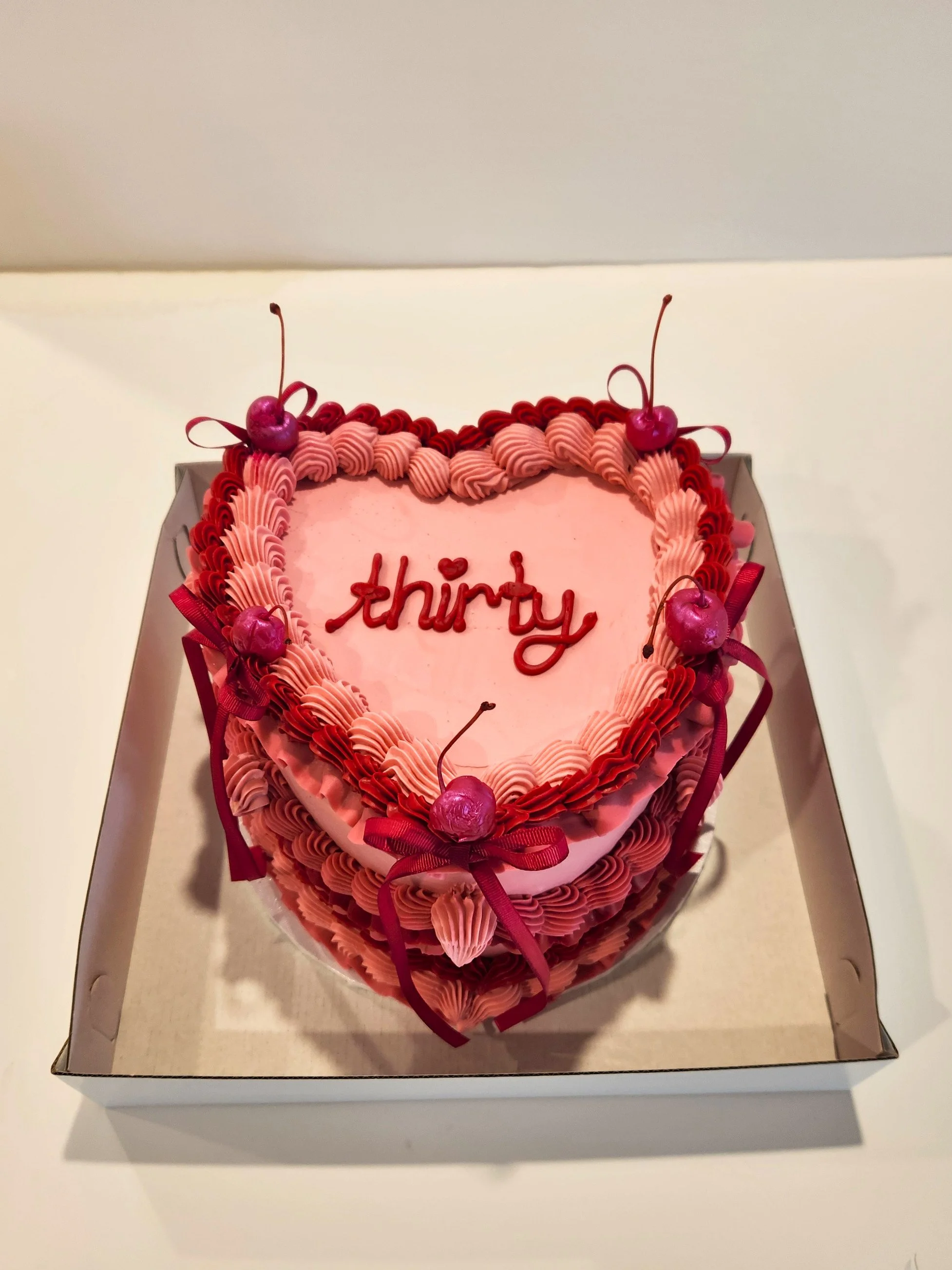 A heart-shaped pink cake with red and pink decorative frosting, topped with glitter cherries and red ribbons, inside a white box.