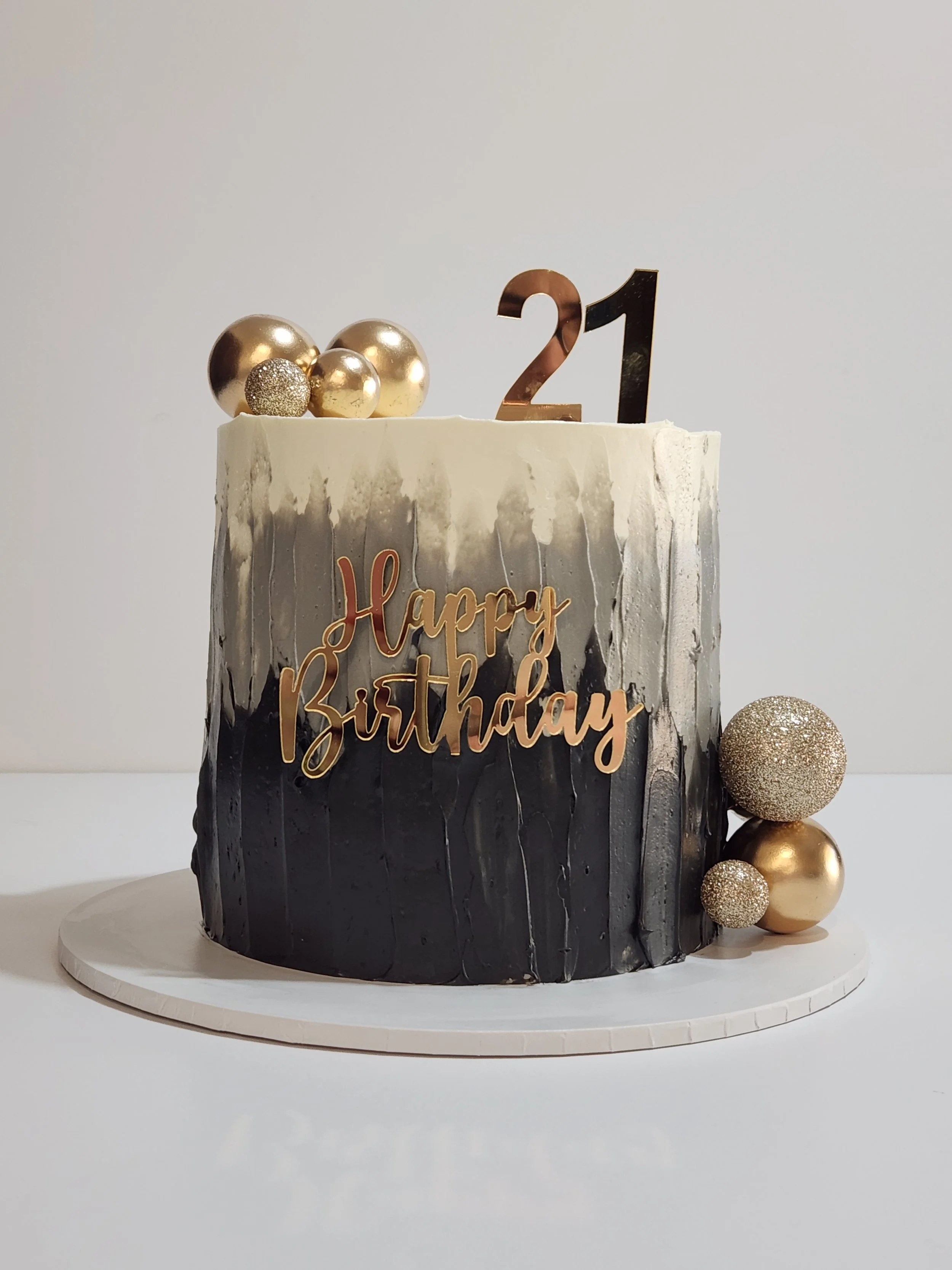 A birthday cake with black and white marble design, gold and silver decorative spheres on top, a gold 'Happy Birthday' sign, and a large gold and glittery sphere to the side, set on a white cake board against a plain background.