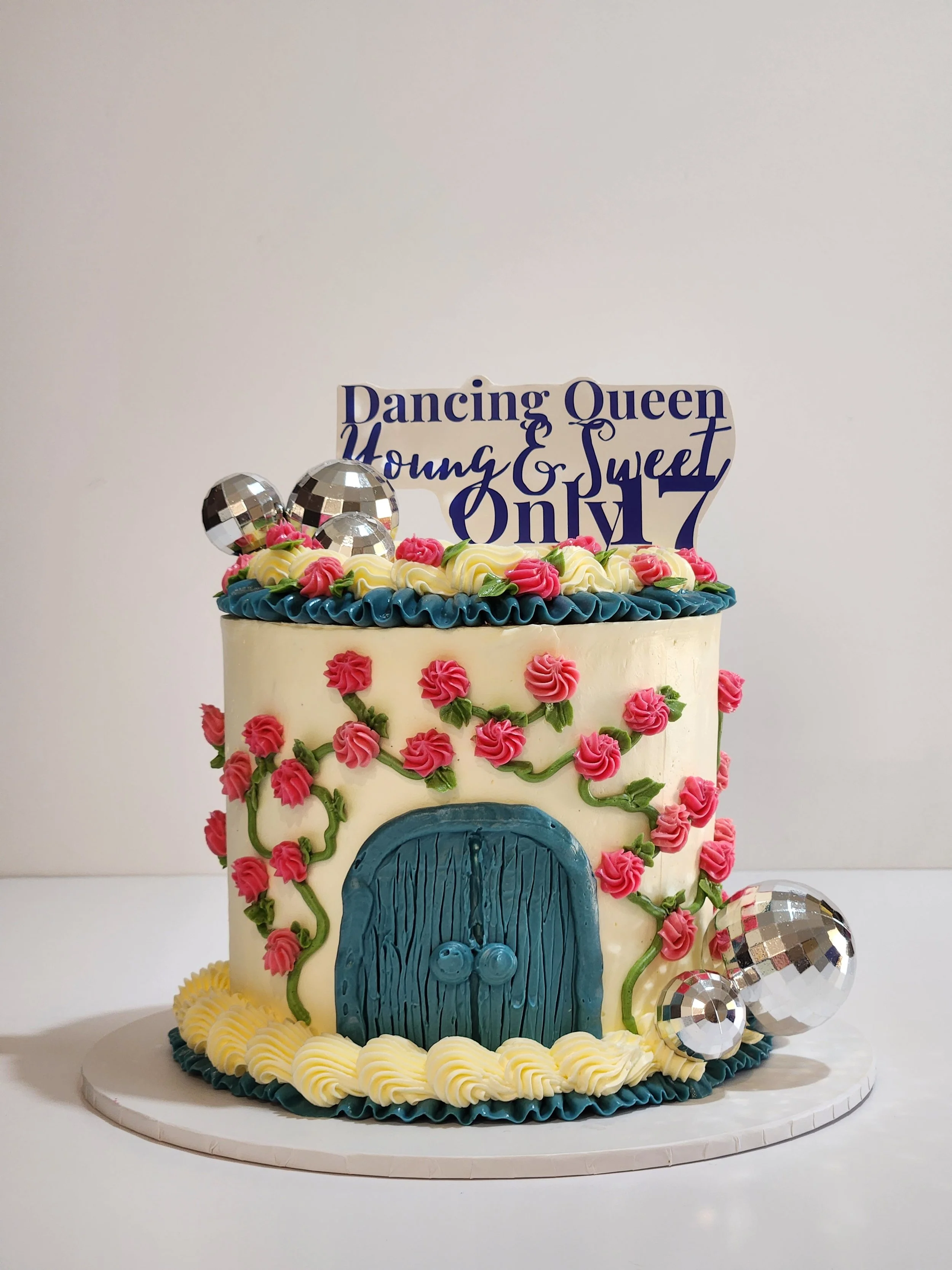 A two-tier birthday cake decorated with pink roses, green vines, blue door, and a sign that reads 'Dancing Queen, Young & Sweet, Only 17.' The cake is adorned with silver disco ball decorations and has a yellow, blue, and white piped frosting border 