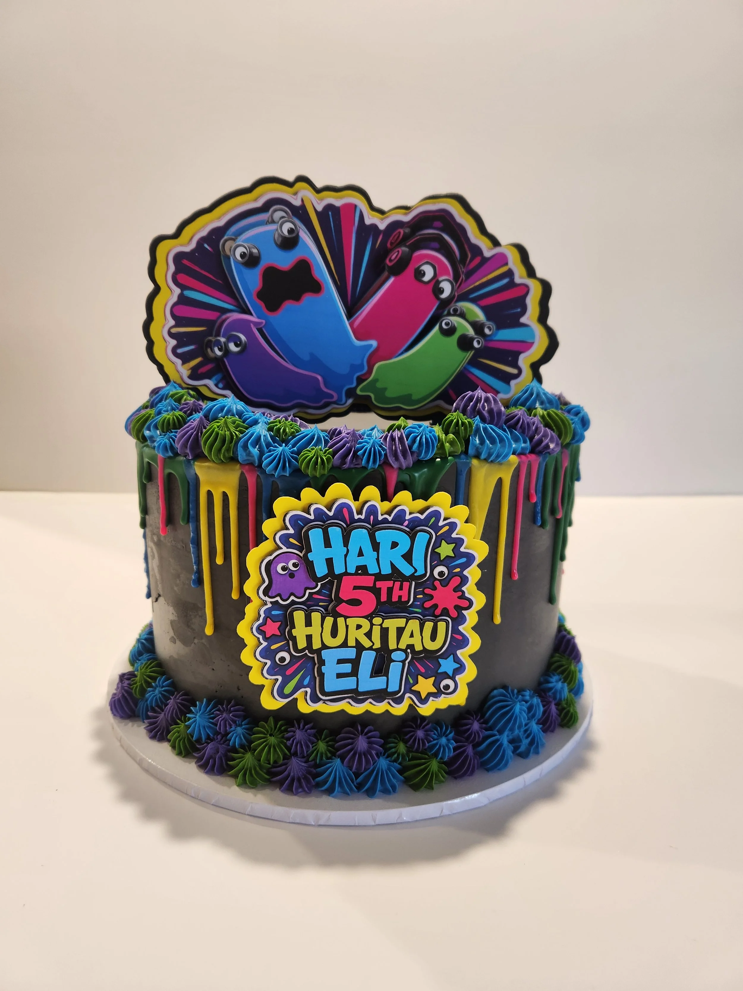 Colorful birthday cake with a 'Happy 5th' message, decorated with purple, green, blue, and pink icing, and a Monster Inc-themed topper featuring Mike and Sulley in vibrant colors.